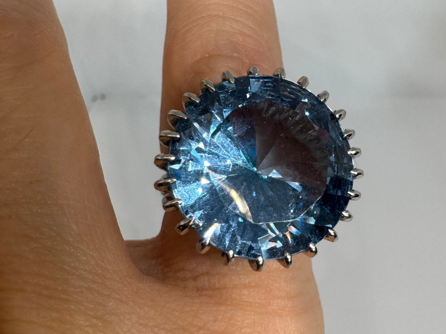 טבעת זהב לבן 14 קראט משובצת אבן טופז בצבע כחול, בולטת, בחיתוך עגול, משובצת במסגרת שיניים גבוהה ליצירת מראה מרשים ותופס עין