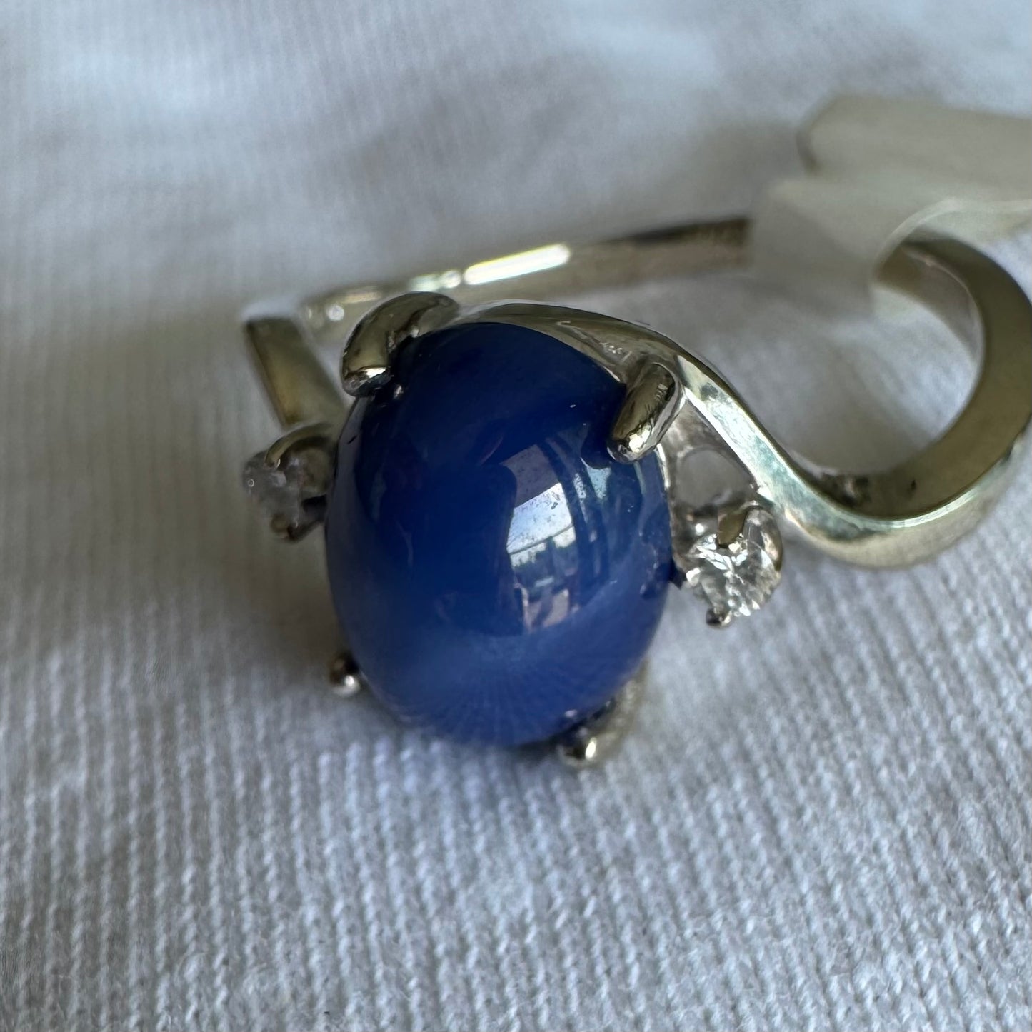 טבעת זהב לבן 14 קראט משובצת אבן ספיר כוכב מרהיבה, המציגה תופעת טבע של שישה קווים זוהרים, ובצידיה שני יהלומים מנצנצים.