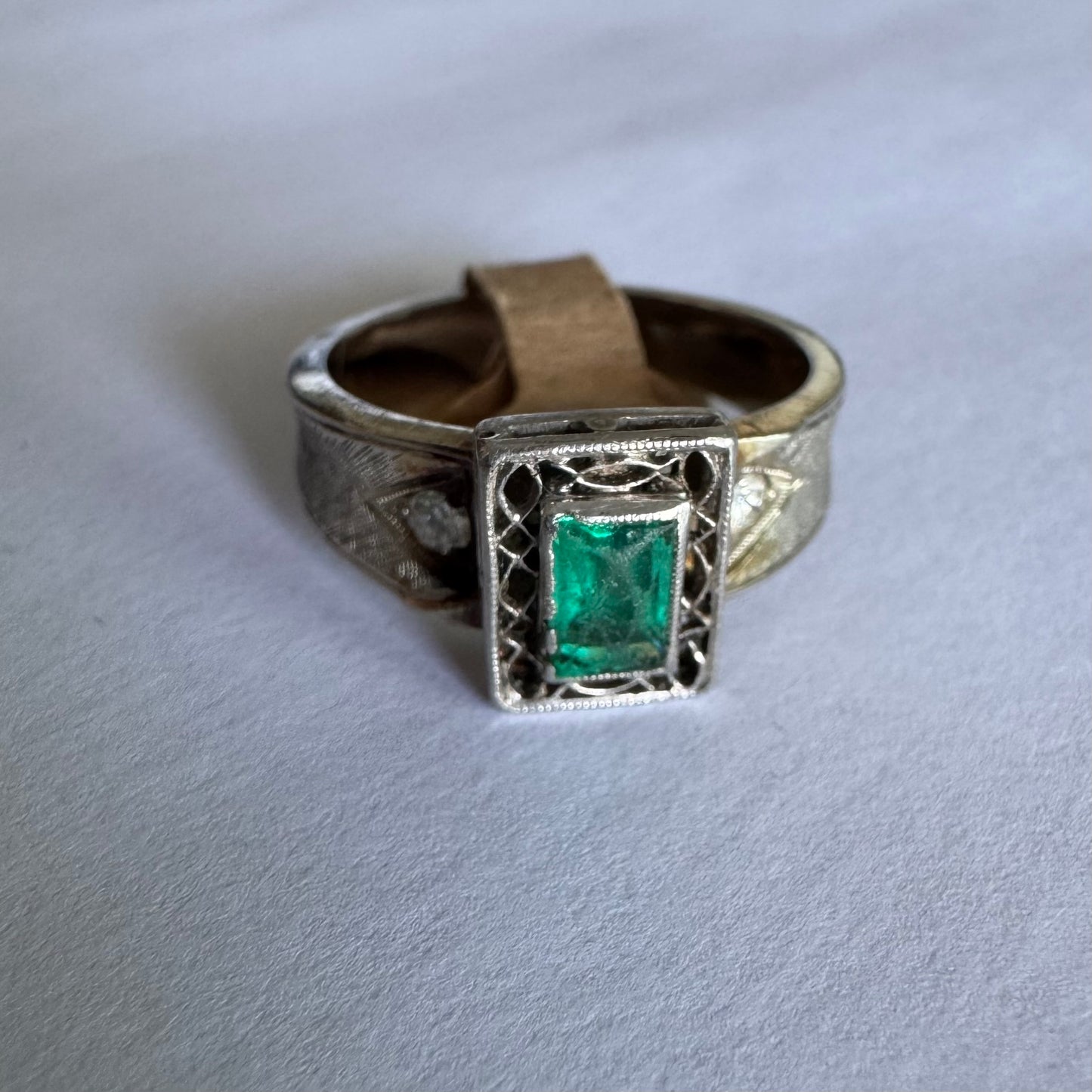 טבעת זהב לבן 14 קראט משובצת אבן אמרלד בחיתוך מלבני (Emerald cut) במרכז, מוקפת במסגרת מלבנית עם עבודת פיליגרן עדינה.