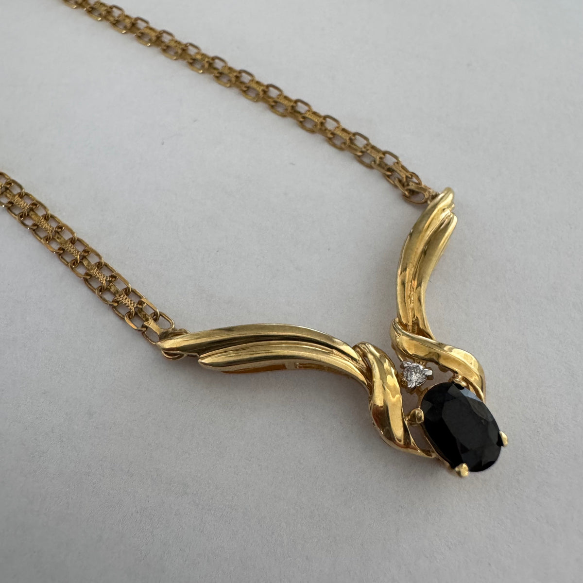 14K gold necklace featuring a flowing pendant design, set with a dark blue center Sapphire and a small Diamond above.