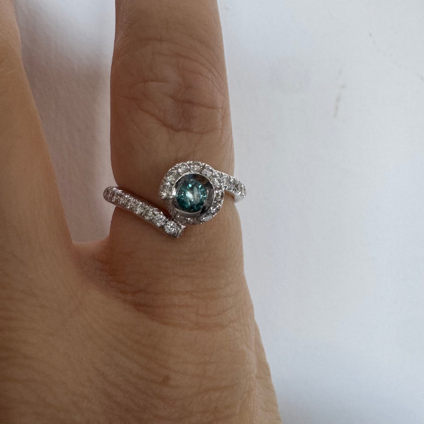 טבעת זהב לבן 18 במרכזה משובץ יהלום כחול עז, מוקף בהילה של יהלומים לבנים והזרועות של הטבעת משובצות יהלומים