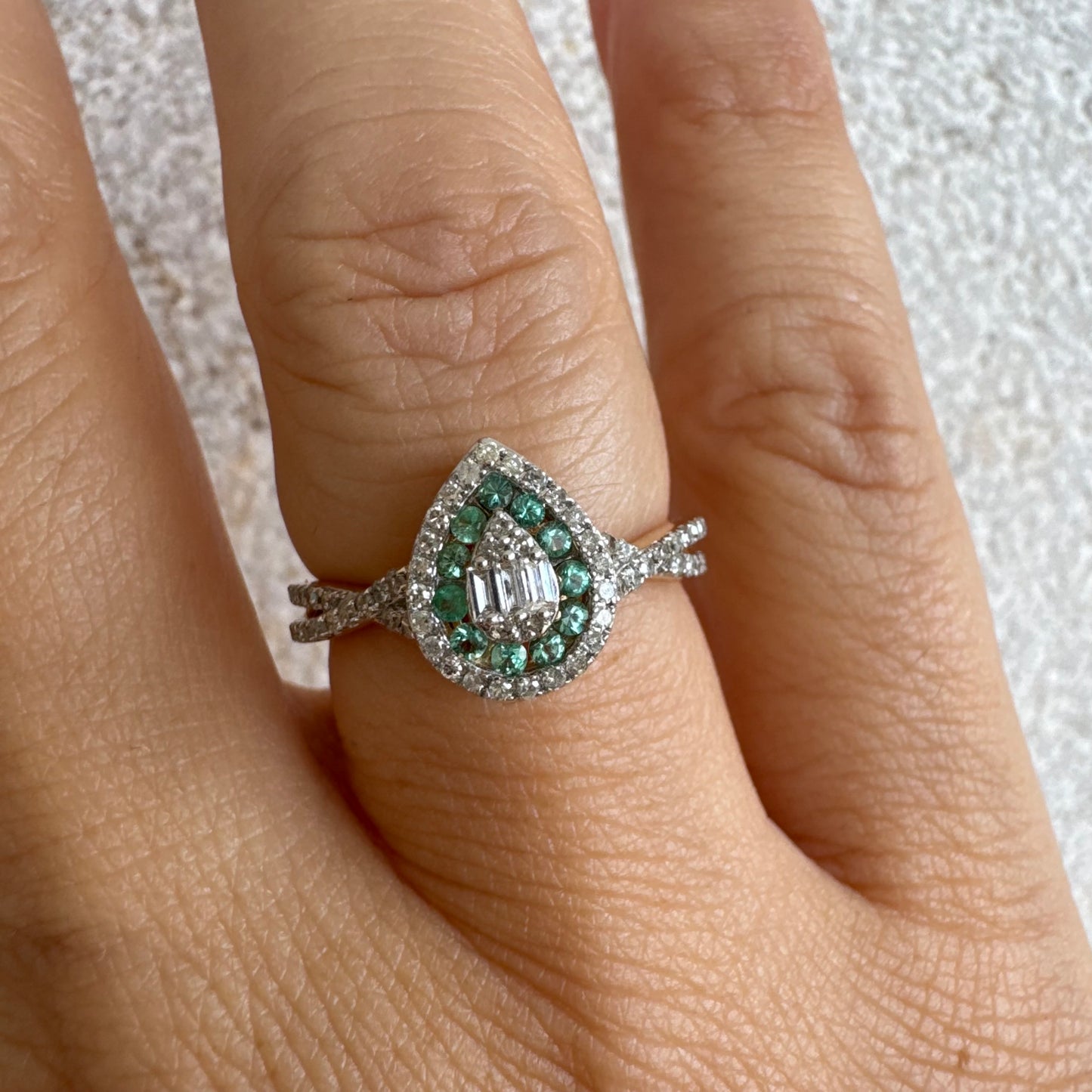 טבעת זהב צהוב 14 קראט בעיצוב טיפה (pear shape), משובצת יהלומים לבנים בשיבוץ הילה כפולה (double halo) ואבני אמרלד קטנות המקיפות מרכז ייחודי של יהלומים בחיתוך באגט