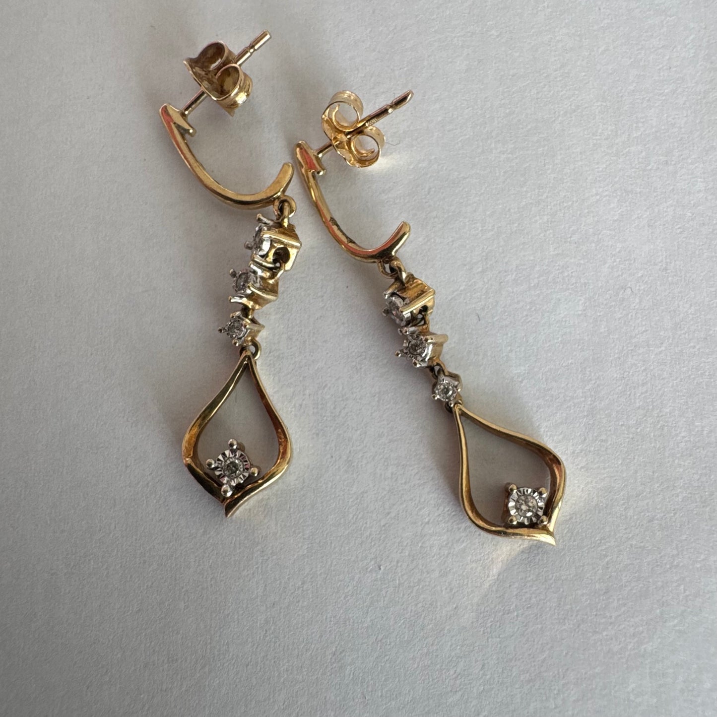 עגילי זהב 14 קראט תלויים בעיצוב עדין ומודרני, משובצים יהלומים לבנים לאורך החלק המרכזי ובטיפה פתוחה בתחתית משובץ יהלום נוסף.