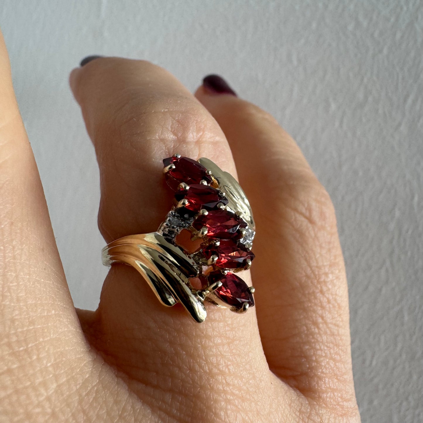 טבעת זהב צהוב 10 קראט בעיצוב דינמי ומלא תנועה, משובצת אבני גרנט בחיתוך מרקיזה בגוון אדום עמוק ובמרכזה נגיעות יהלומים לבנים.