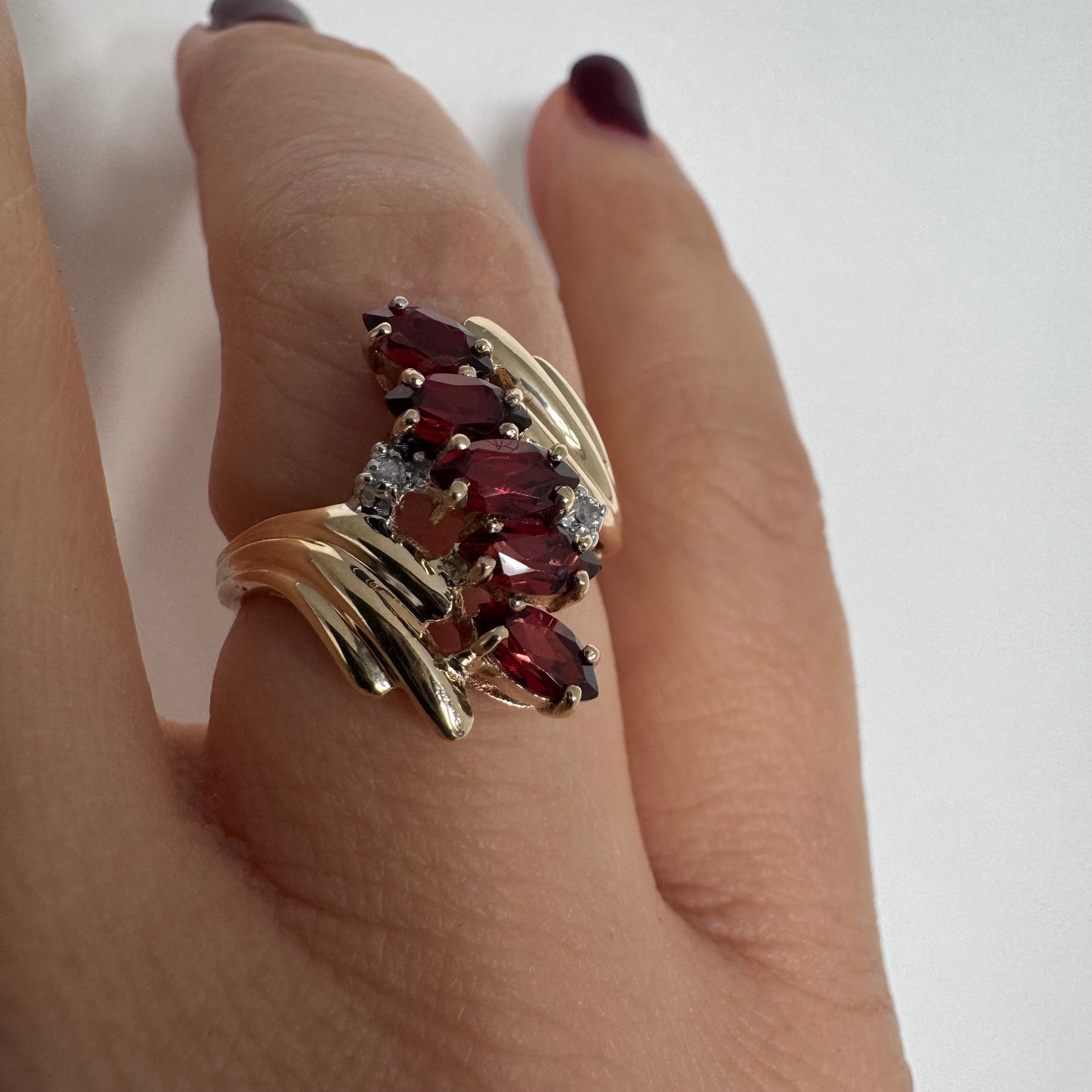 טבעת זהב צהוב 10 קראט בעיצוב דינמי ומלא תנועה, משובצת אבני גרנט בחיתוך מרקיזה בגוון אדום עמוק ובמרכזה נגיעות יהלומים לבנים.