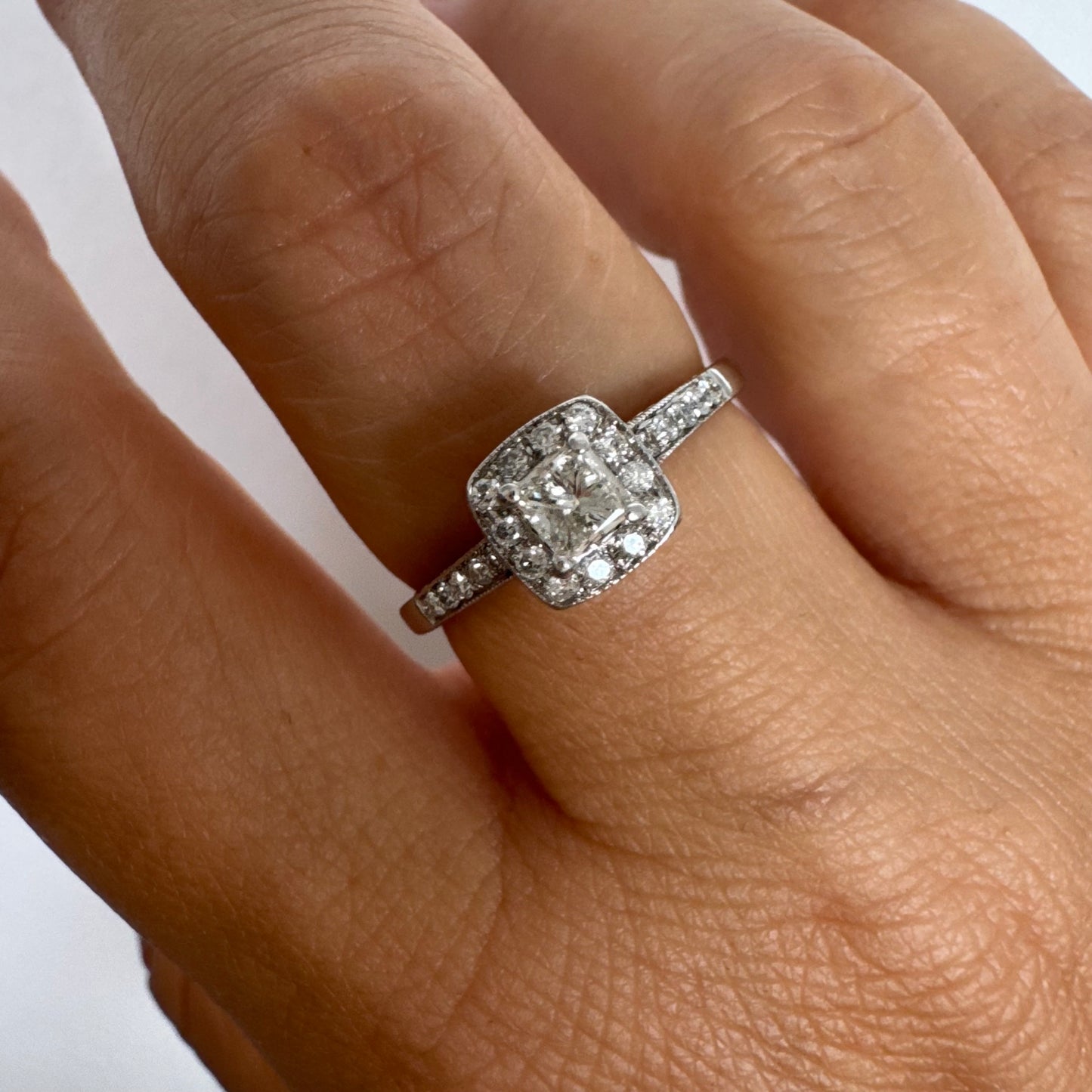 טבעת זהב לבן 14 קראט משובצת יהלום מרכזי בחיתוך פרינסס, מוקף הילת יהלומים קטנים וזרוע משובצת פאווה