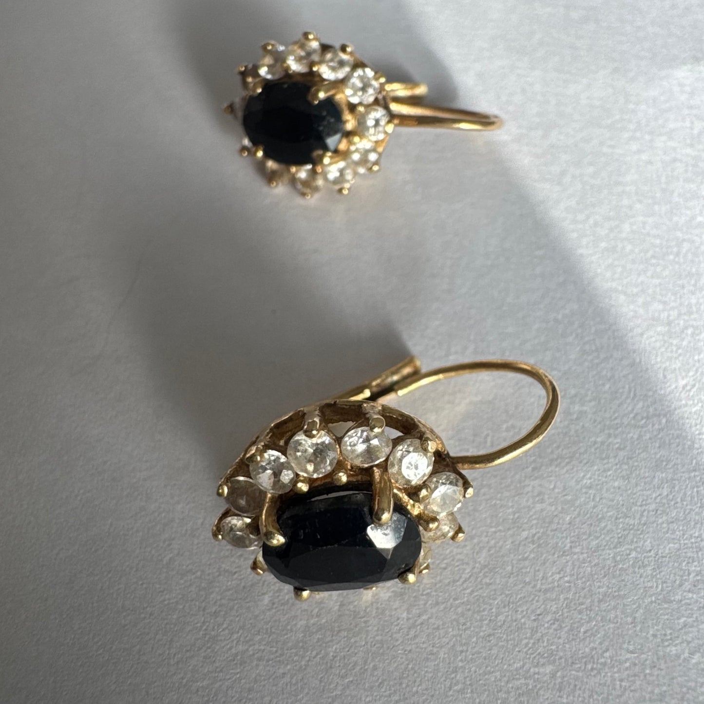 עגילי זהב צהוב 14 קראט בעיצוב קלאסי ומרשים, משובצים אבן ספיר בגוון כחול כהה בחיתוך אובלי, ומסביבה משובצים אבנים נוצצות שמוסיפות אור וזוהר לכל תנועה.