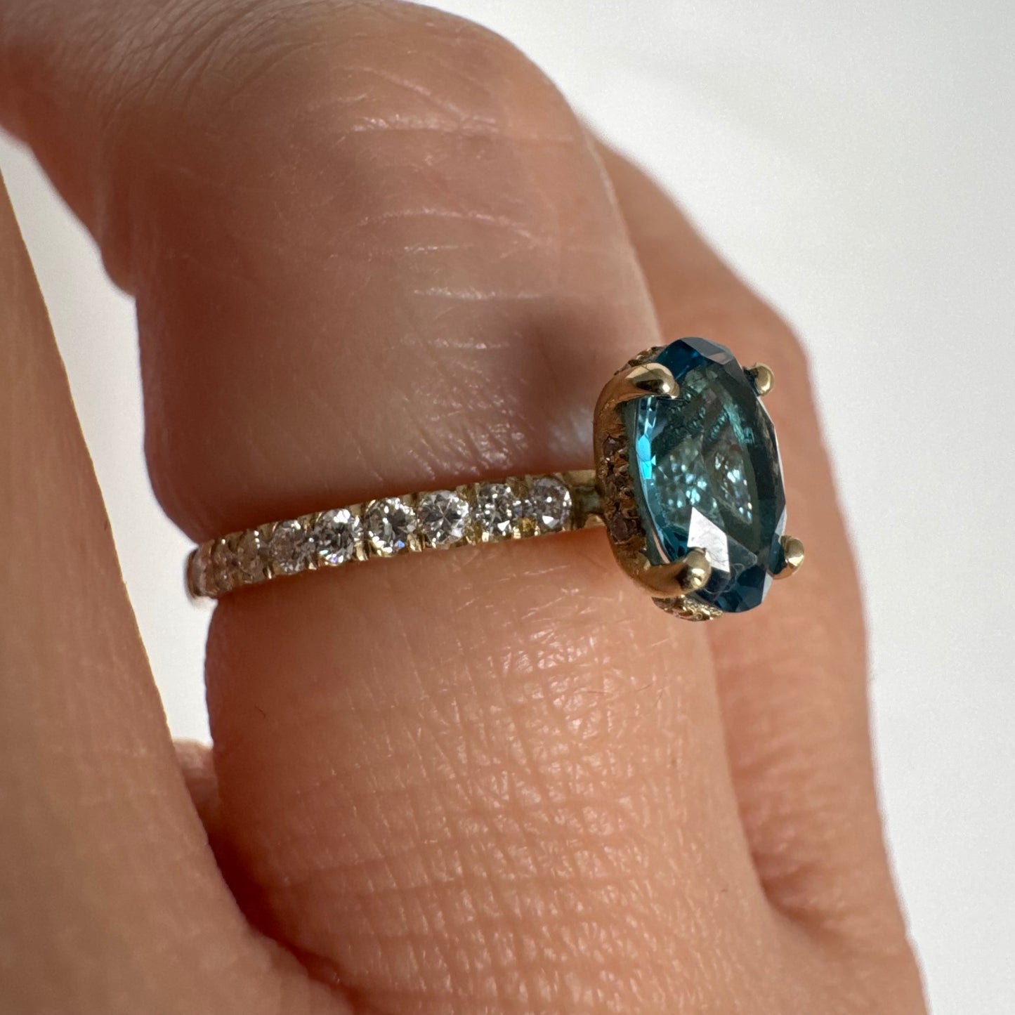טבעת זהב צהוב 14 קראט משובצת אבן טופז כחולה אובלית בגוון טורקיז־כחול עמוק, באחיזה של ארבע שיניים עדינות ויהלומים.