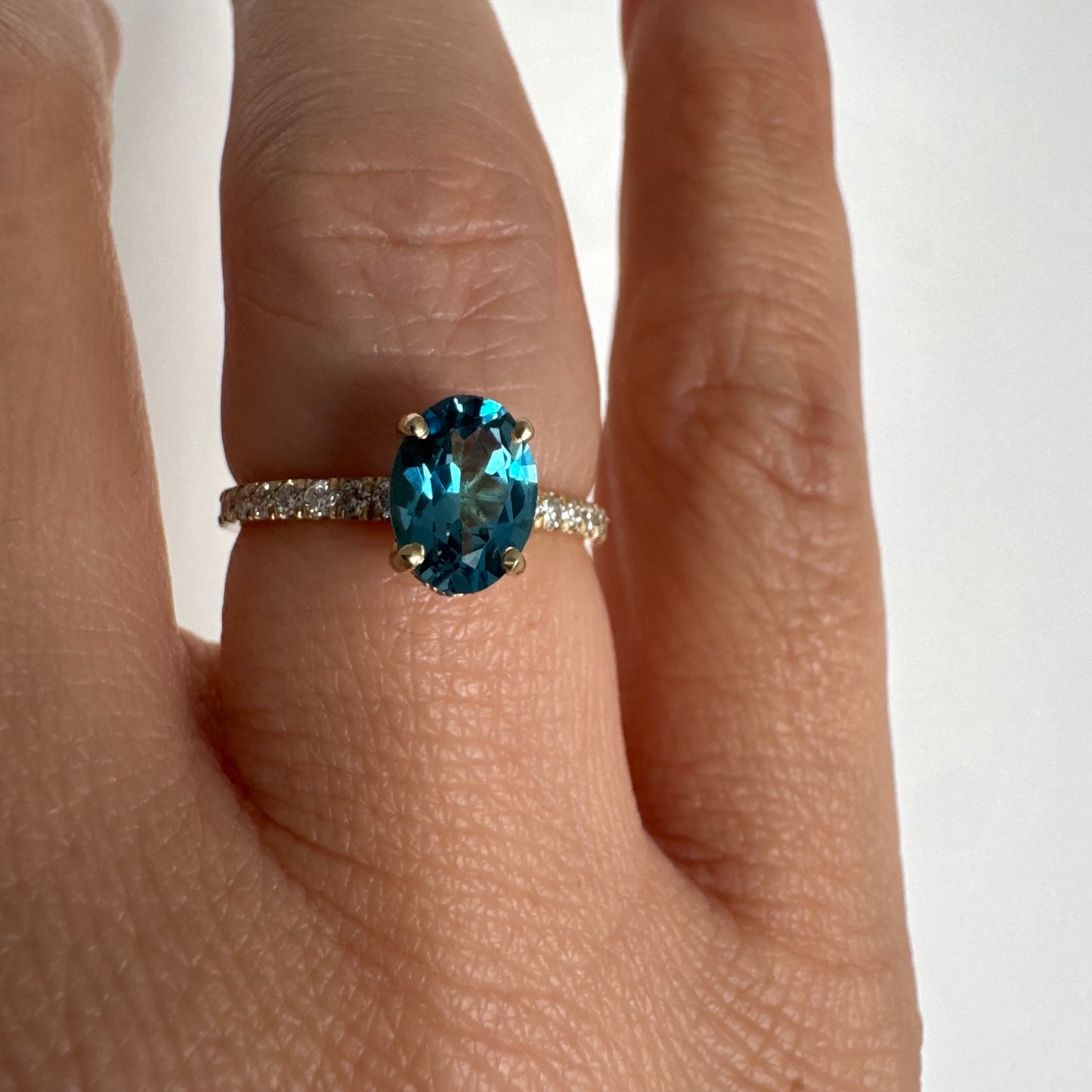 טבעת זהב צהוב 14 קראט משובצת אבן טופז כחולה אובלית בגוון טורקיז־כחול עמוק, באחיזה של ארבע שיניים עדינות ויהלומים.