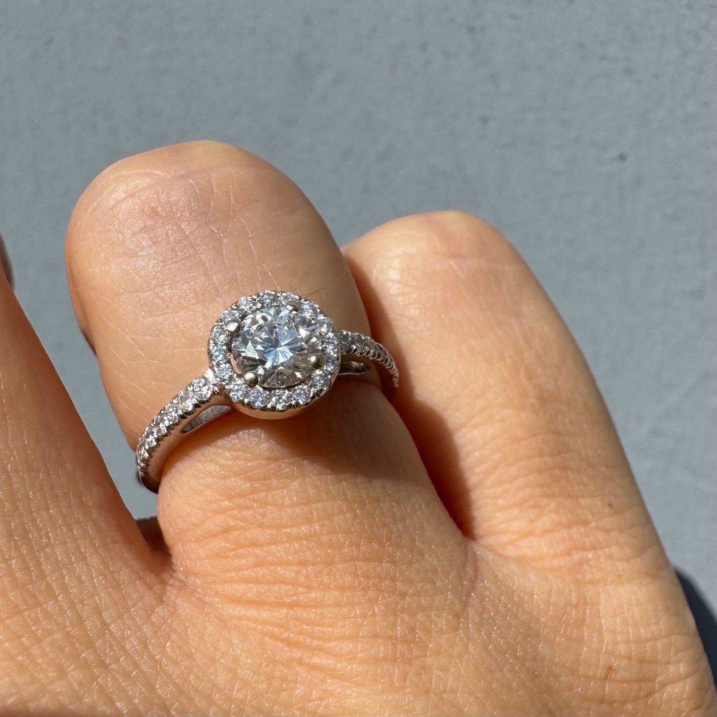 טבעת זהב לבן 14 קראט משובצת יהלום מרכזי עגול והילה נוצצת של יהלומים קטנים, עם יהלומים נוספים על החישוק.