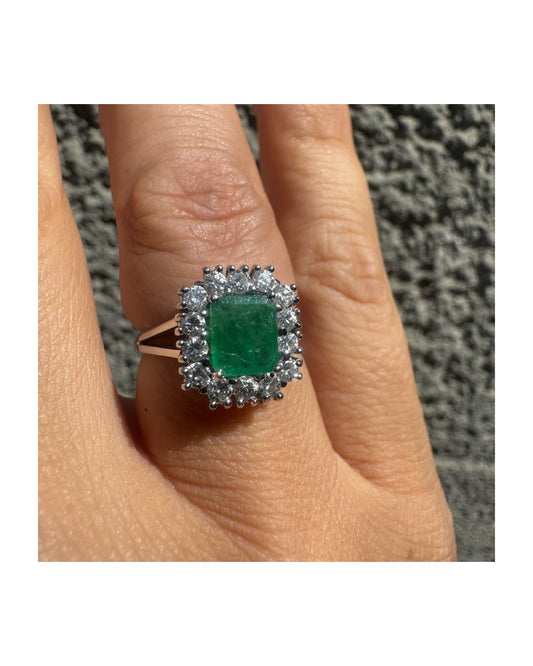 18K gold ring set with a green Emerald at the center, surrounded by a halo of sparkling round Diamonds.