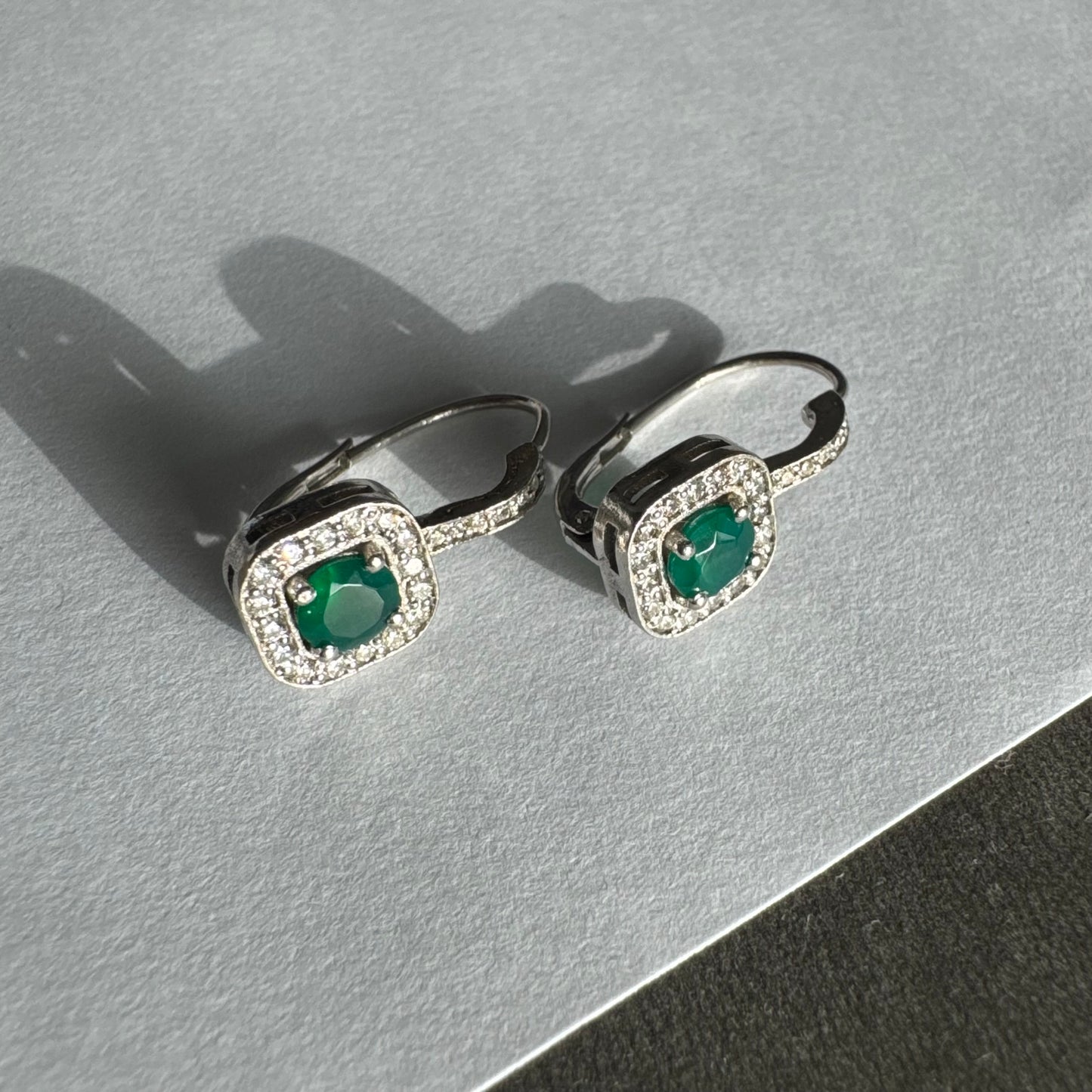 עגילי זהב לבן 14 קראט בעיצוב אלגנטי ונקי, משובצים אבן אמרלד מרכזית במראה עשיר ועמוק, מוקפת הילה מנצנצת של יהלומים.