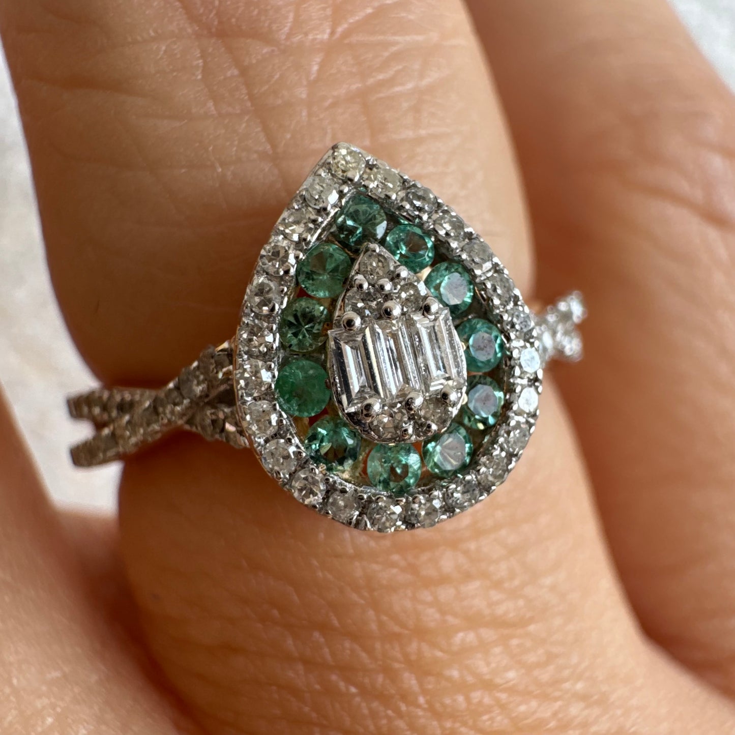 טבעת זהב צהוב 14 קראט בעיצוב טיפה (pear shape), משובצת יהלומים לבנים בשיבוץ הילה כפולה (double halo) ואבני אמרלד קטנות המקיפות מרכז ייחודי של יהלומים בחיתוך באגט