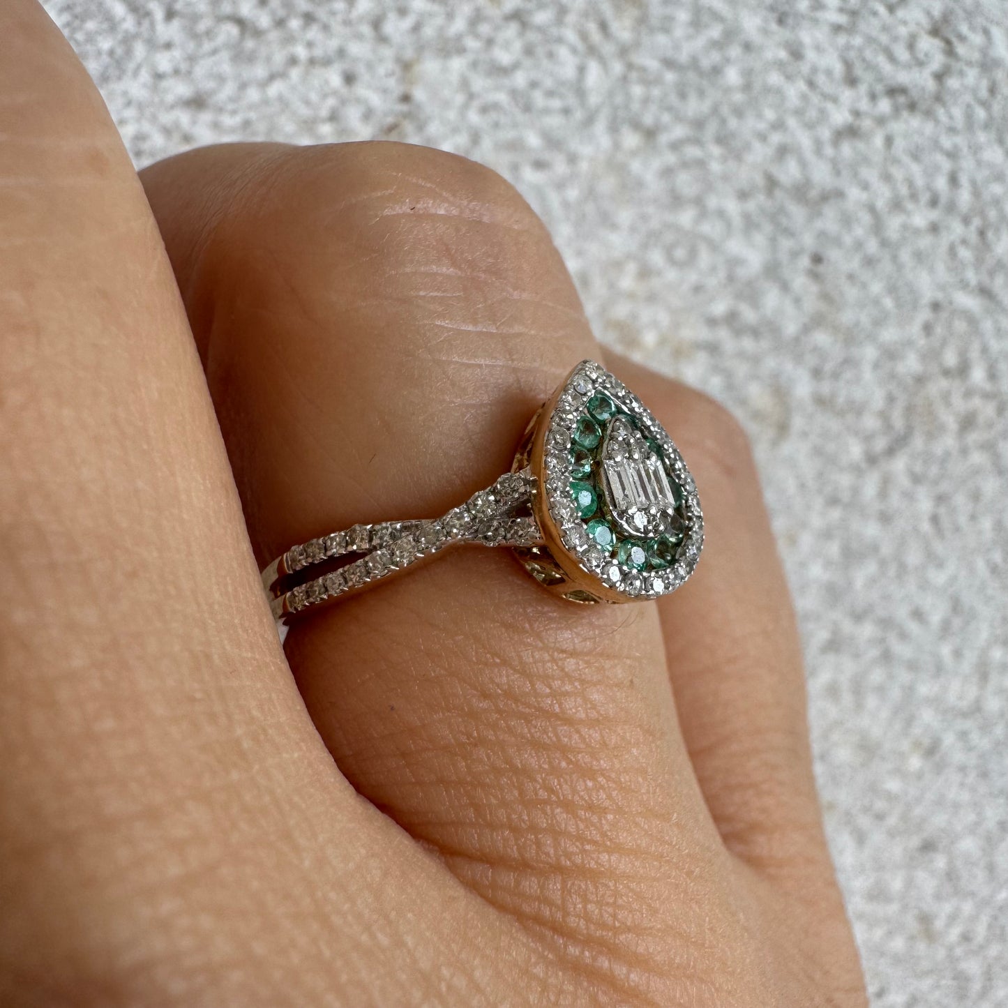 טבעת זהב צהוב 14 קראט בעיצוב טיפה (pear shape), משובצת יהלומים לבנים בשיבוץ הילה כפולה (double halo) ואבני אמרלד קטנות המקיפות מרכז ייחודי של יהלומים בחיתוך באגט