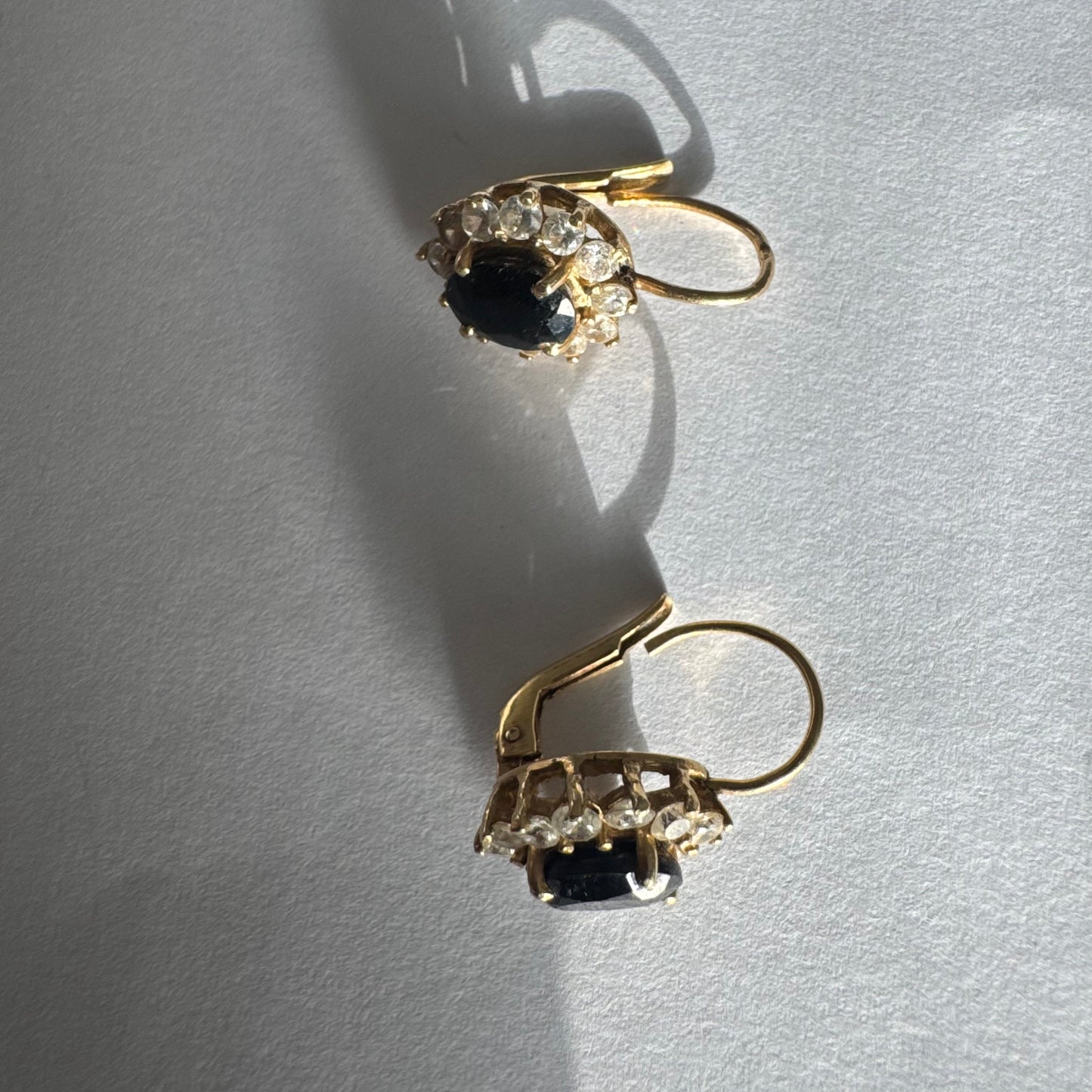 עגילי זהב צהוב 14 קראט בעיצוב קלאסי ומרשים, משובצים אבן ספיר בגוון כחול כהה בחיתוך אובלי, ומסביבה משובצים אבנים נוצצות שמוסיפות אור וזוהר לכל תנועה.