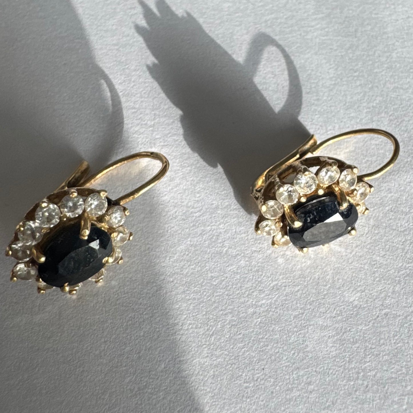 עגילי זהב צהוב 14 קראט בעיצוב קלאסי ומרשים, משובצים אבן ספיר בגוון כחול כהה בחיתוך אובלי, ומסביבה משובצים אבנים נוצצות שמוסיפות אור וזוהר לכל תנועה.