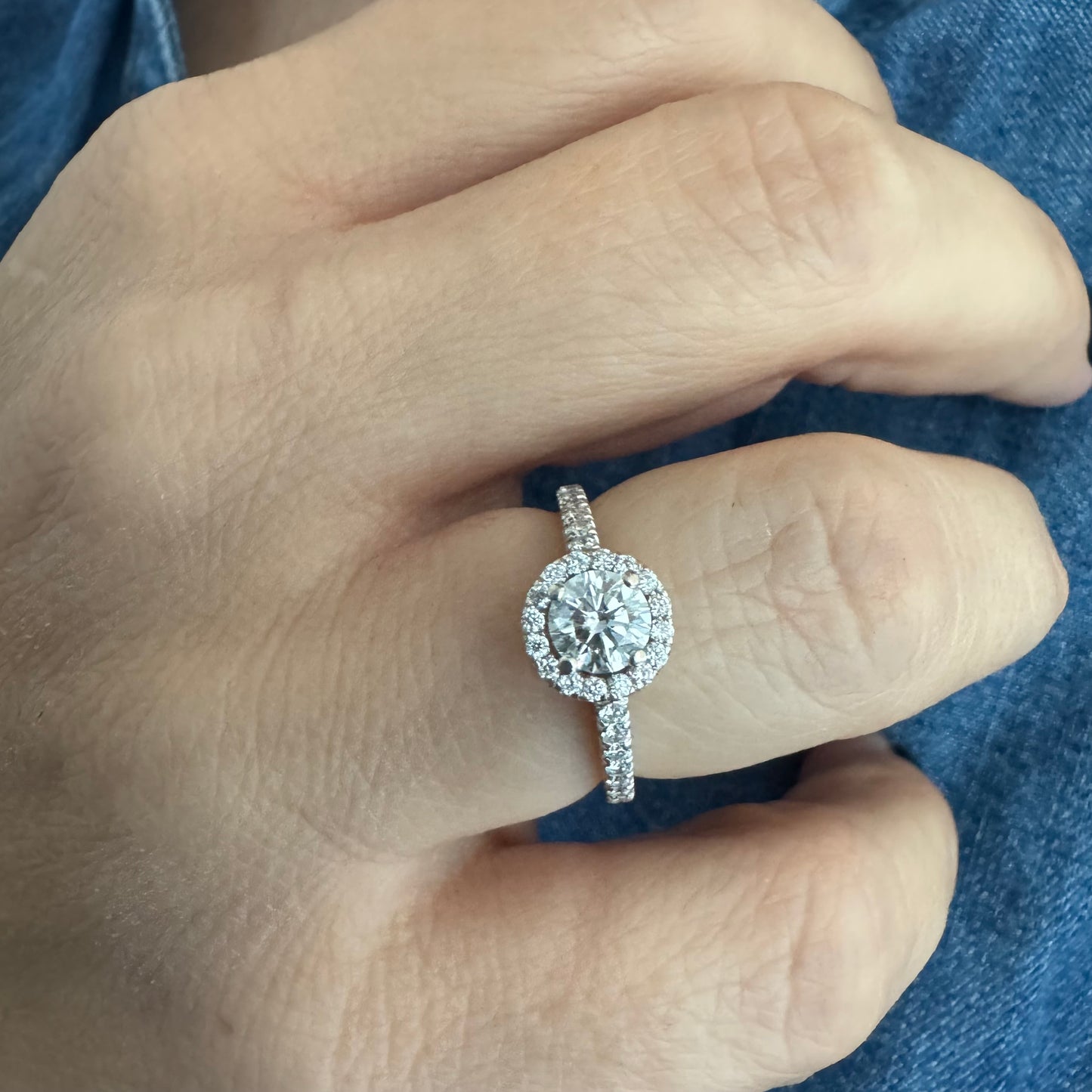 טבעת זהב לבן 14 קראט משובצת יהלום מרכזי עגול והילה נוצצת של יהלומים קטנים, עם יהלומים נוספים על החישוק.