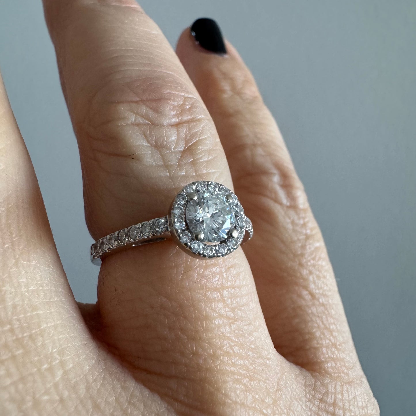 טבעת זהב לבן 14 קראט משובצת יהלום מרכזי עגול והילה נוצצת של יהלומים קטנים, עם יהלומים נוספים על החישוק.