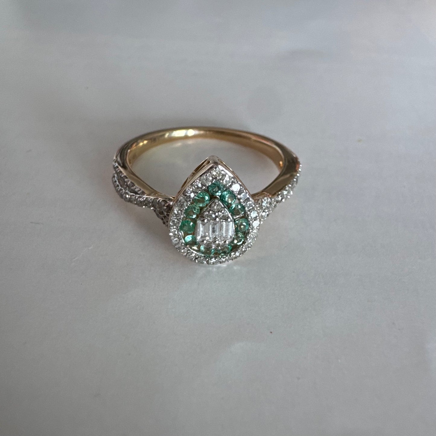 טבעת זהב צהוב 14 קראט בעיצוב טיפה (pear shape), משובצת יהלומים לבנים בשיבוץ הילה כפולה (double halo) ואבני אמרלד קטנות המקיפות מרכז ייחודי של יהלומים בחיתוך באגט