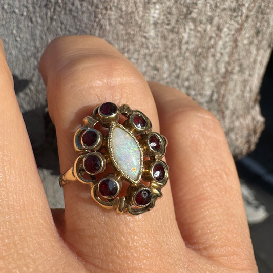 טבעת זהב צהוב 14 קראט משובצת באבן אופל מרכזית בחיתוך מרקיזה, מוקפת באבני גרנט עגולות כהות.