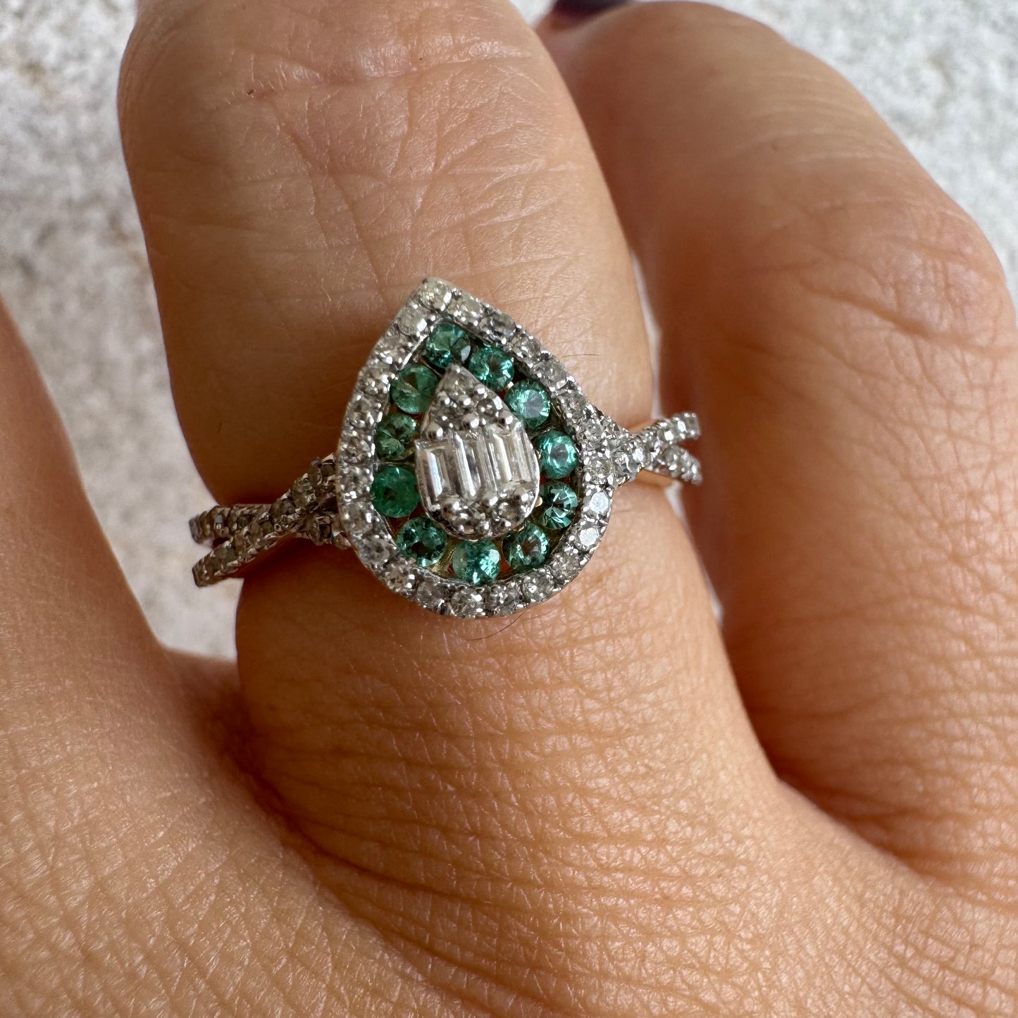 טבעת זהב צהוב 14 קראט בעיצוב טיפה (pear shape), משובצת יהלומים לבנים בשיבוץ הילה כפולה (double halo) ואבני אמרלד קטנות המקיפות מרכז ייחודי של יהלומים בחיתוך באגט