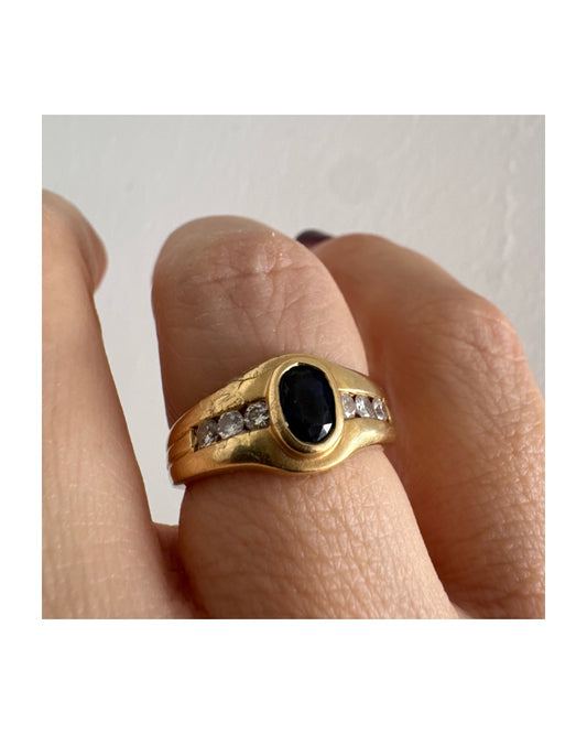 טבעת זהב צהוב 18 קראט משובצת אבן ספיר אובלית בגוון כחול עמוק, במסגרת חלקה ומודגשת, ומשני צדדיה שורת יהלומים משובצים בתעלה.