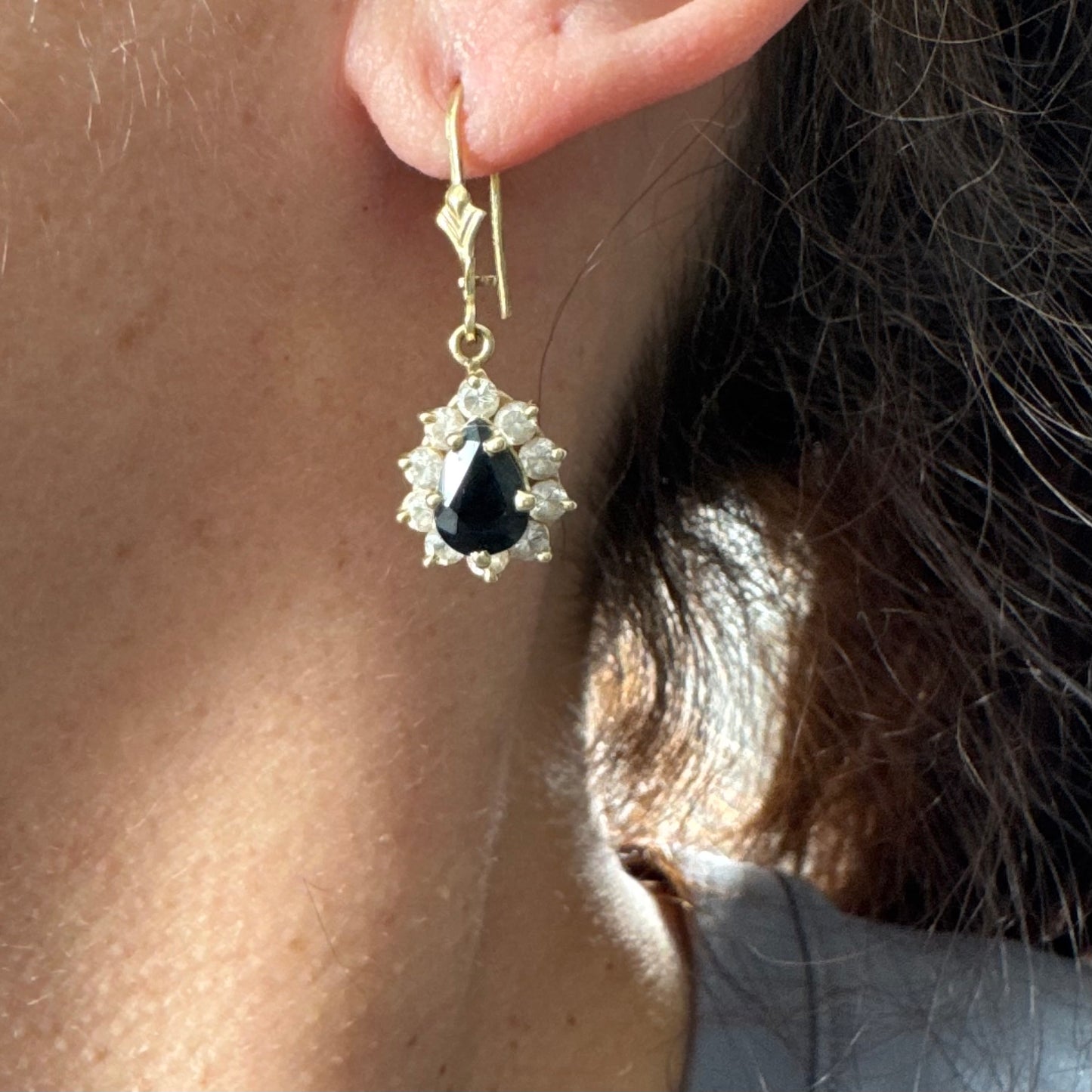 עגילי זהב צהוב 14 קראט בעיצוב קלאסי ומרשים, משובצים אבן ספיר בגוון כחול כהה בחיתוך טיפה ומסביבה אבנים שקופות עם הילה נוצצת שמוסיפה אור וזוהר לכל תנועה.