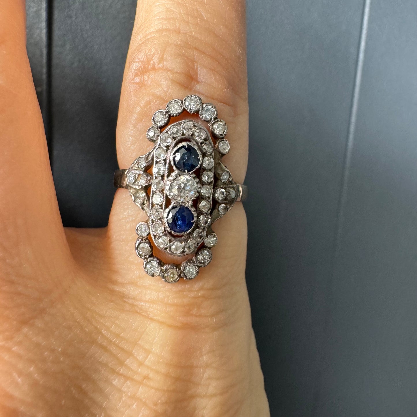 טבעת זהב לבן 14 קראט עתיקה עוצרת נשימה, בעיצוב מאורך ואלגנטי, המשובצת בשלישיית אבנים — שתי אבני ספיר בצבע כחול עמוק ובאמצע משובץ יהלום מרכזי בחיתוך עתיק.