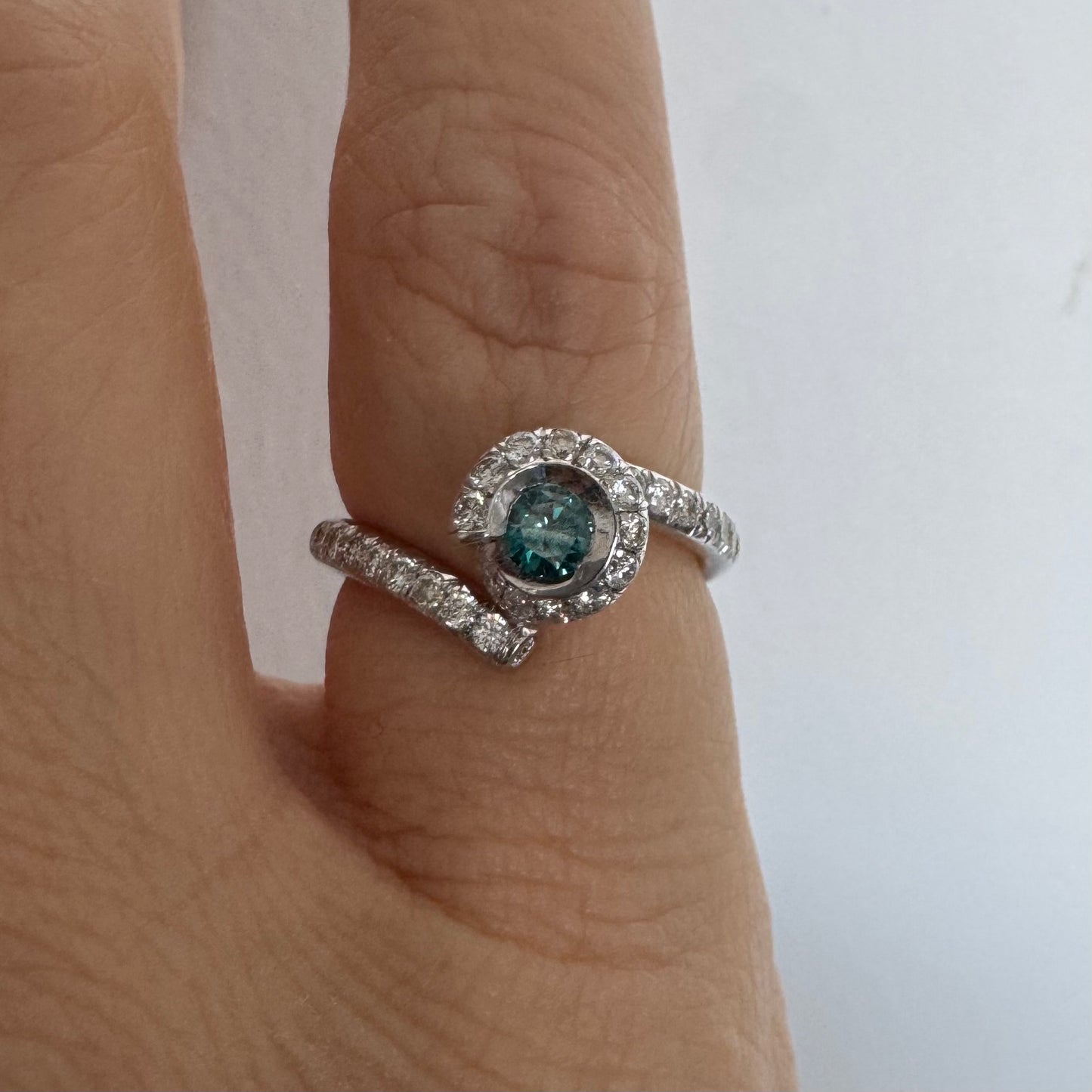 טבעת זהב לבן 18 במרכזה משובץ יהלום כחול עז, מוקף בהילה של יהלומים לבנים והזרועות של הטבעת משובצות יהלומים