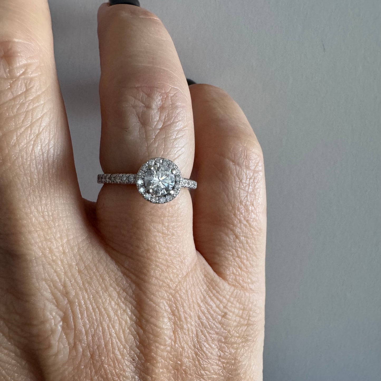 טבעת זהב לבן 14 קראט משובצת יהלום מרכזי עגול והילה נוצצת של יהלומים קטנים, עם יהלומים נוספים על החישוק.