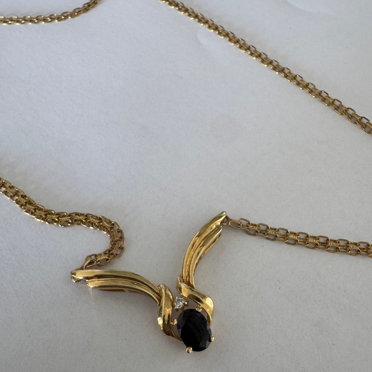 14K gold necklace featuring a flowing pendant design, set with a dark blue center Sapphire and a small Diamond above.
