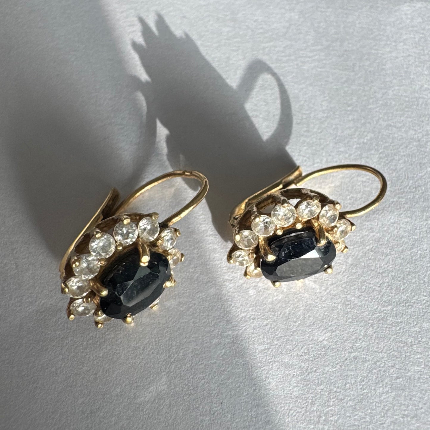 עגילי זהב צהוב 14 קראט בעיצוב קלאסי ומרשים, משובצים אבן ספיר בגוון כחול כהה בחיתוך אובלי, ומסביבה משובצים אבנים נוצצות שמוסיפות אור וזוהר לכל תנועה.