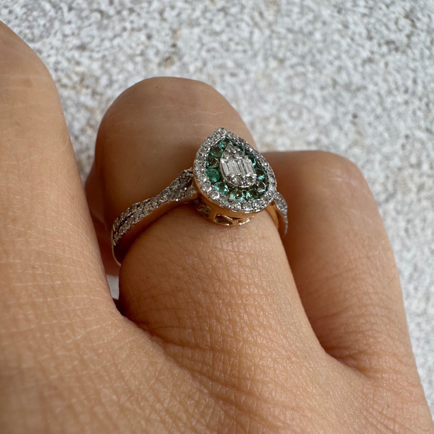 טבעת זהב צהוב 14 קראט בעיצוב טיפה (pear shape), משובצת יהלומים לבנים בשיבוץ הילה כפולה (double halo) ואבני אמרלד קטנות המקיפות מרכז ייחודי של יהלומים בחיתוך באגט