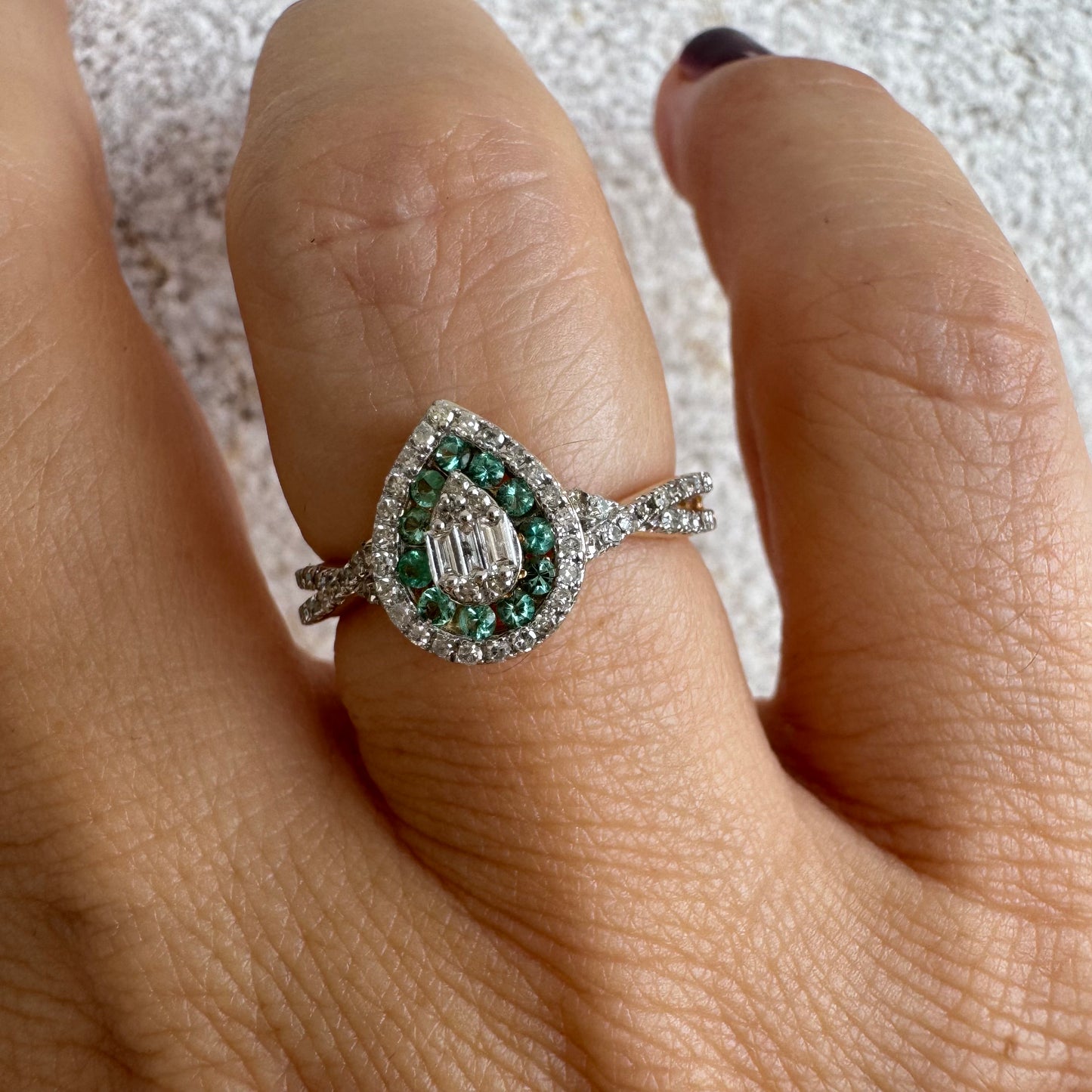 טבעת זהב צהוב 14 קראט בעיצוב טיפה (pear shape), משובצת יהלומים לבנים בשיבוץ הילה כפולה (double halo) ואבני אמרלד קטנות המקיפות מרכז ייחודי של יהלומים בחיתוך באגט