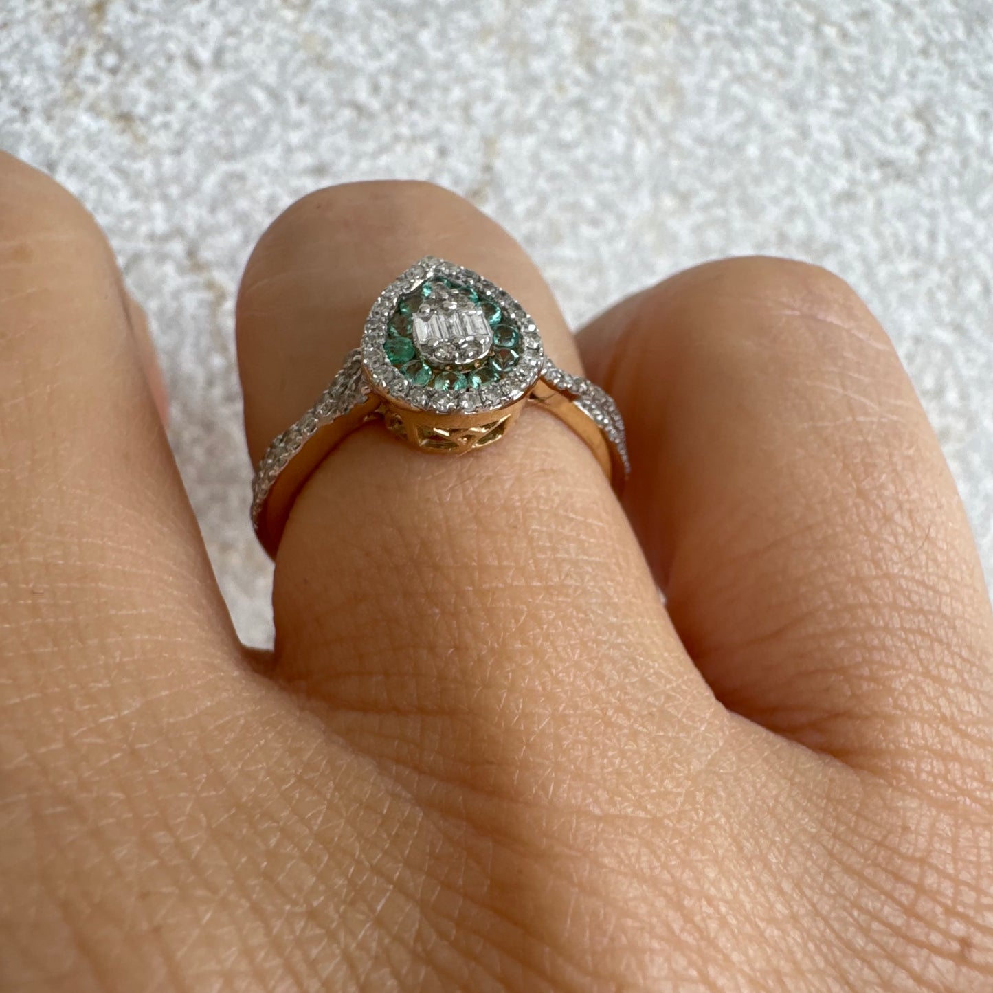 טבעת זהב צהוב 14 קראט בעיצוב טיפה (pear shape), משובצת יהלומים לבנים בשיבוץ הילה כפולה (double halo) ואבני אמרלד קטנות המקיפות מרכז ייחודי של יהלומים בחיתוך באגט