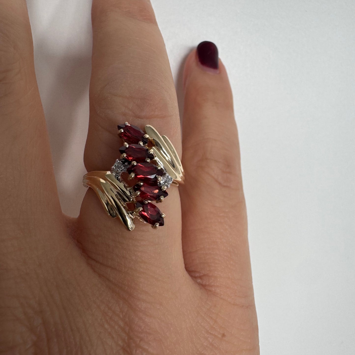 טבעת זהב צהוב 10 קראט בעיצוב דינמי ומלא תנועה, משובצת אבני גרנט בחיתוך מרקיזה בגוון אדום עמוק ובמרכזה נגיעות יהלומים לבנים.