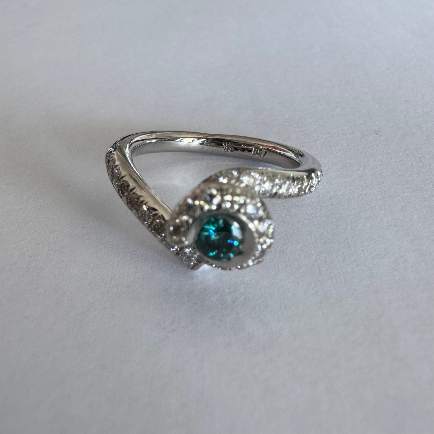 טבעת זהב לבן 18 במרכזה משובץ יהלום כחול עז, מוקף בהילה של יהלומים לבנים והזרועות של הטבעת משובצות יהלומים