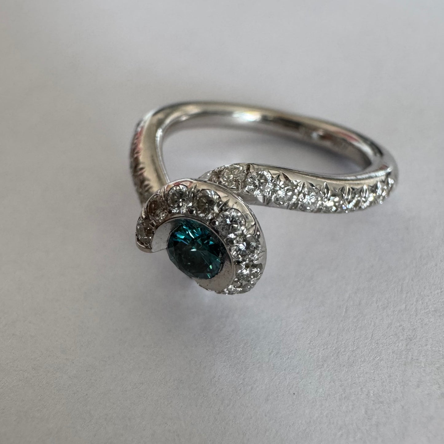 טבעת זהב לבן 18 במרכזה משובץ יהלום כחול עז, מוקף בהילה של יהלומים לבנים והזרועות של הטבעת משובצות יהלומים