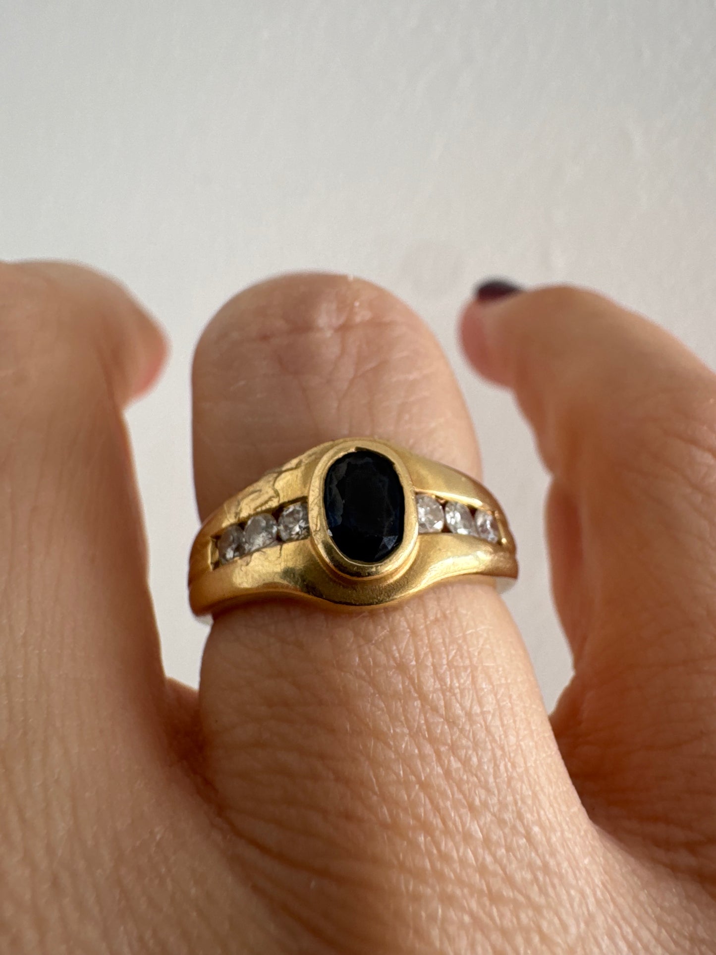 טבעת זהב צהוב 18 קראט משובצת אבן ספיר אובלית בגוון כחול עמוק, במסגרת חלקה ומודגשת, ומשני צדדיה שורת יהלומים משובצים בתעלה.