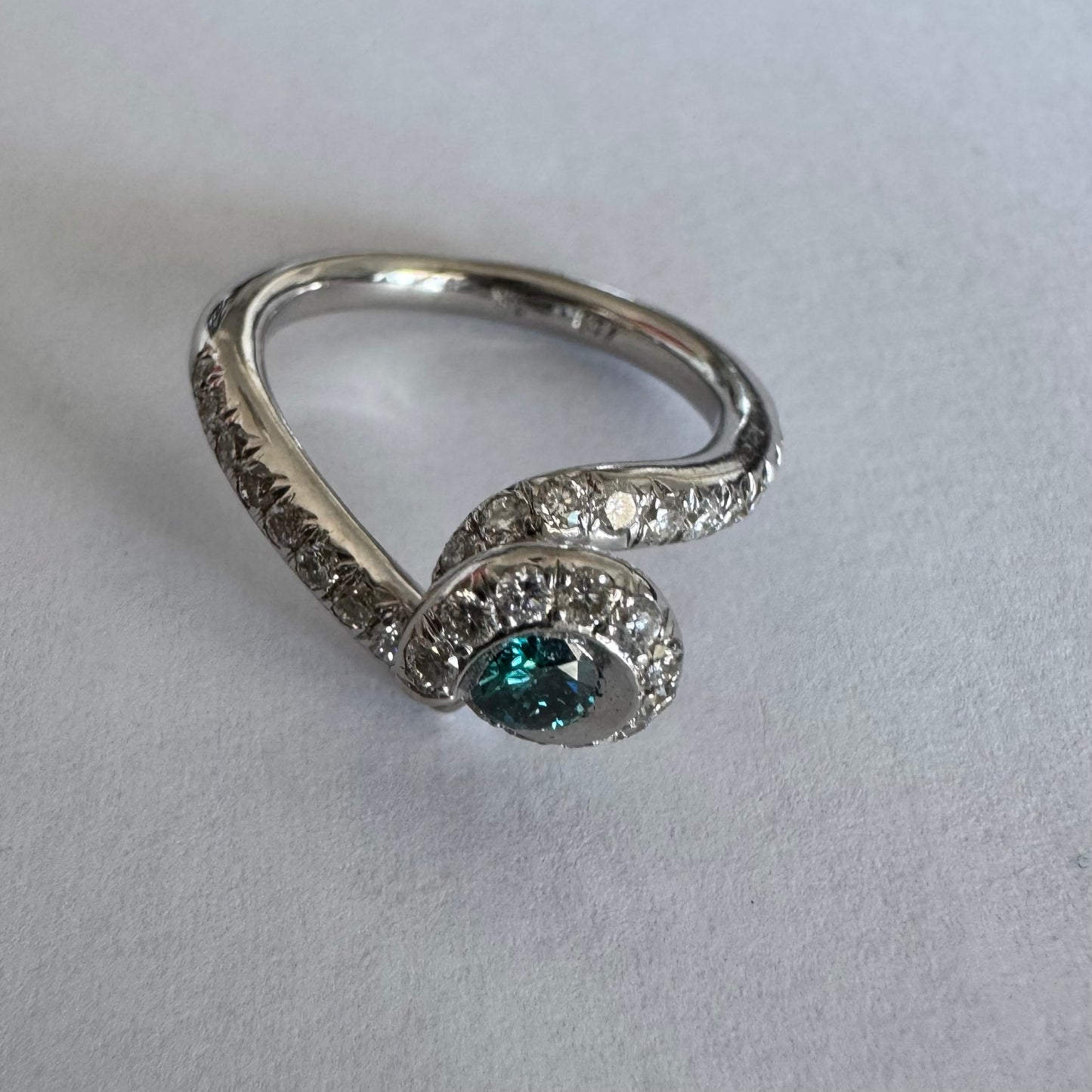 טבעת זהב לבן 18 במרכזה משובץ יהלום כחול עז, מוקף בהילה של יהלומים לבנים והזרועות של הטבעת משובצות יהלומים