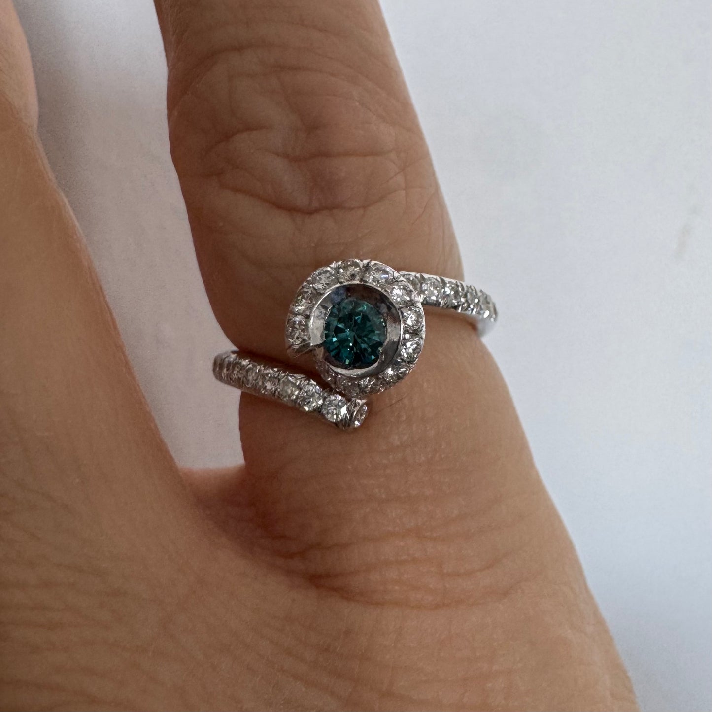 טבעת זהב לבן 18 במרכזה משובץ יהלום כחול עז, מוקף בהילה של יהלומים לבנים והזרועות של הטבעת משובצות יהלומים