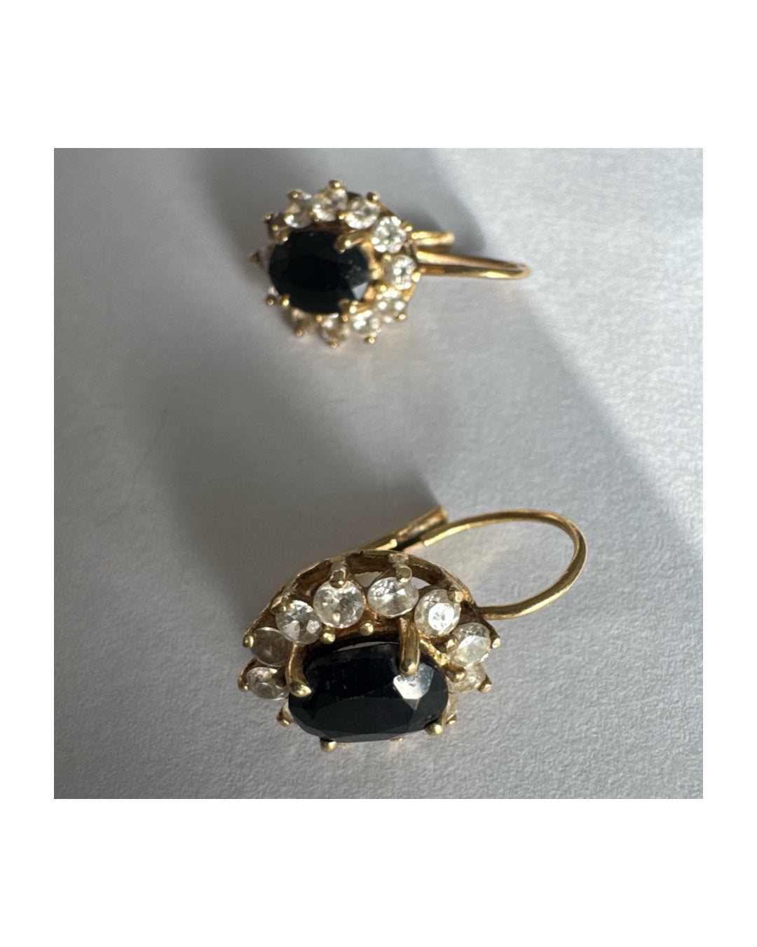 עגילי זהב צהוב 14 קראט בעיצוב קלאסי ומרשים, משובצים אבן ספיר בגוון כחול כהה בחיתוך אובלי, ומסביבה משובצים אבנים נוצצות שמוסיפות אור וזוהר לכל תנועה.