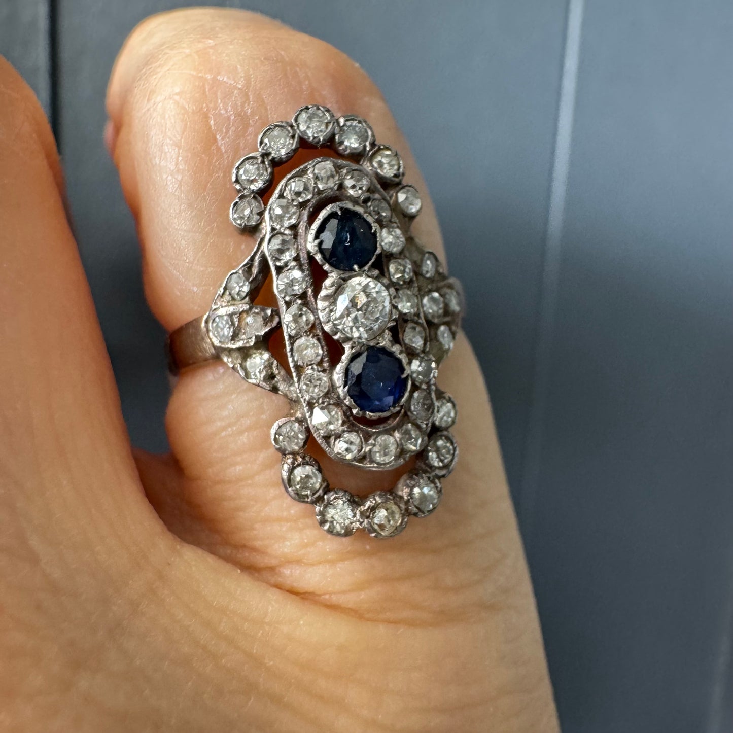 טבעת זהב לבן 14 קראט עתיקה עוצרת נשימה, בעיצוב מאורך ואלגנטי, המשובצת בשלישיית אבנים — שתי אבני ספיר בצבע כחול עמוק ובאמצע משובץ יהלום מרכזי בחיתוך עתיק.