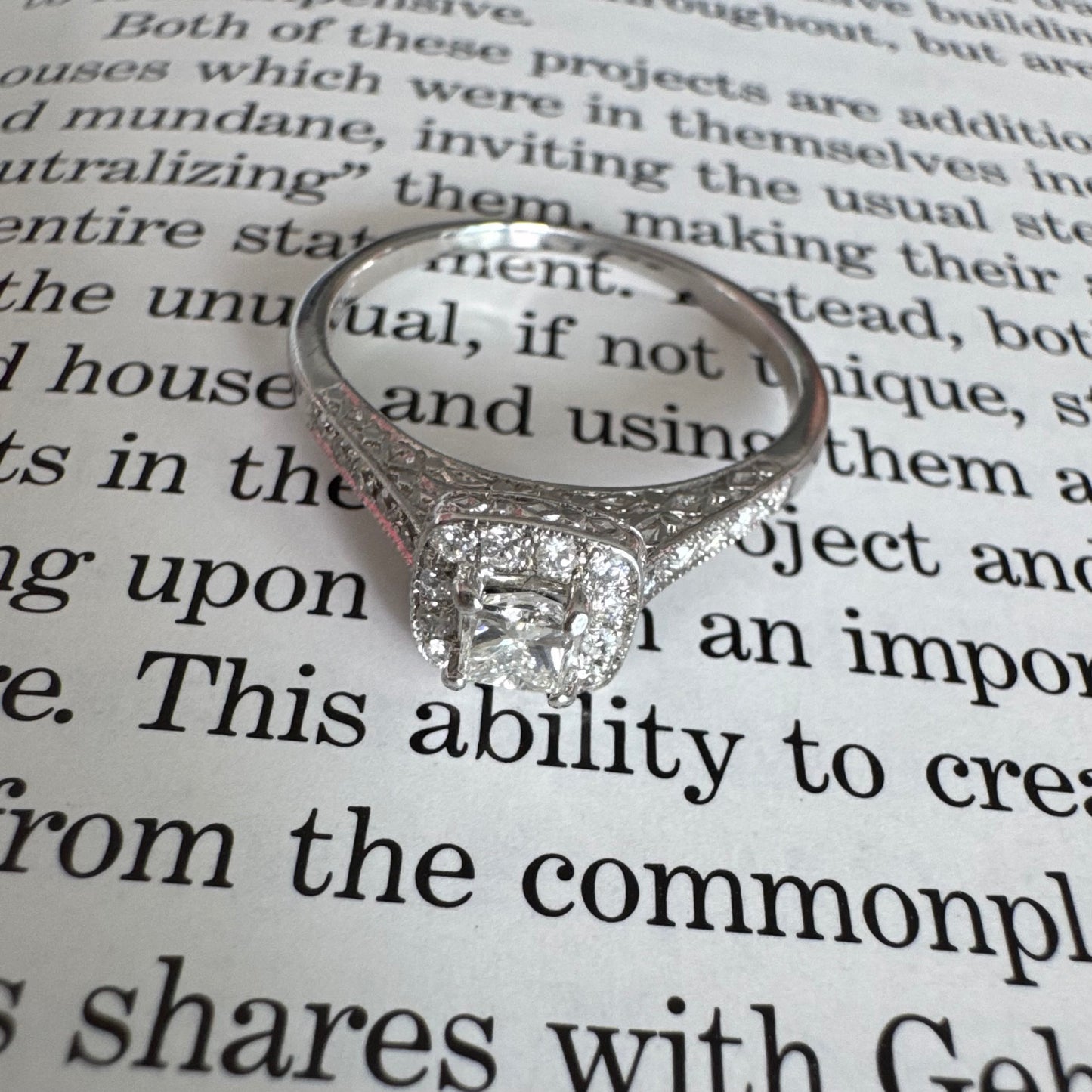 טבעת זהב לבן 14 קראט משובצת יהלום מרכזי בחיתוך פרינסס, מוקף הילת יהלומים קטנים וזרוע משובצת פאווה