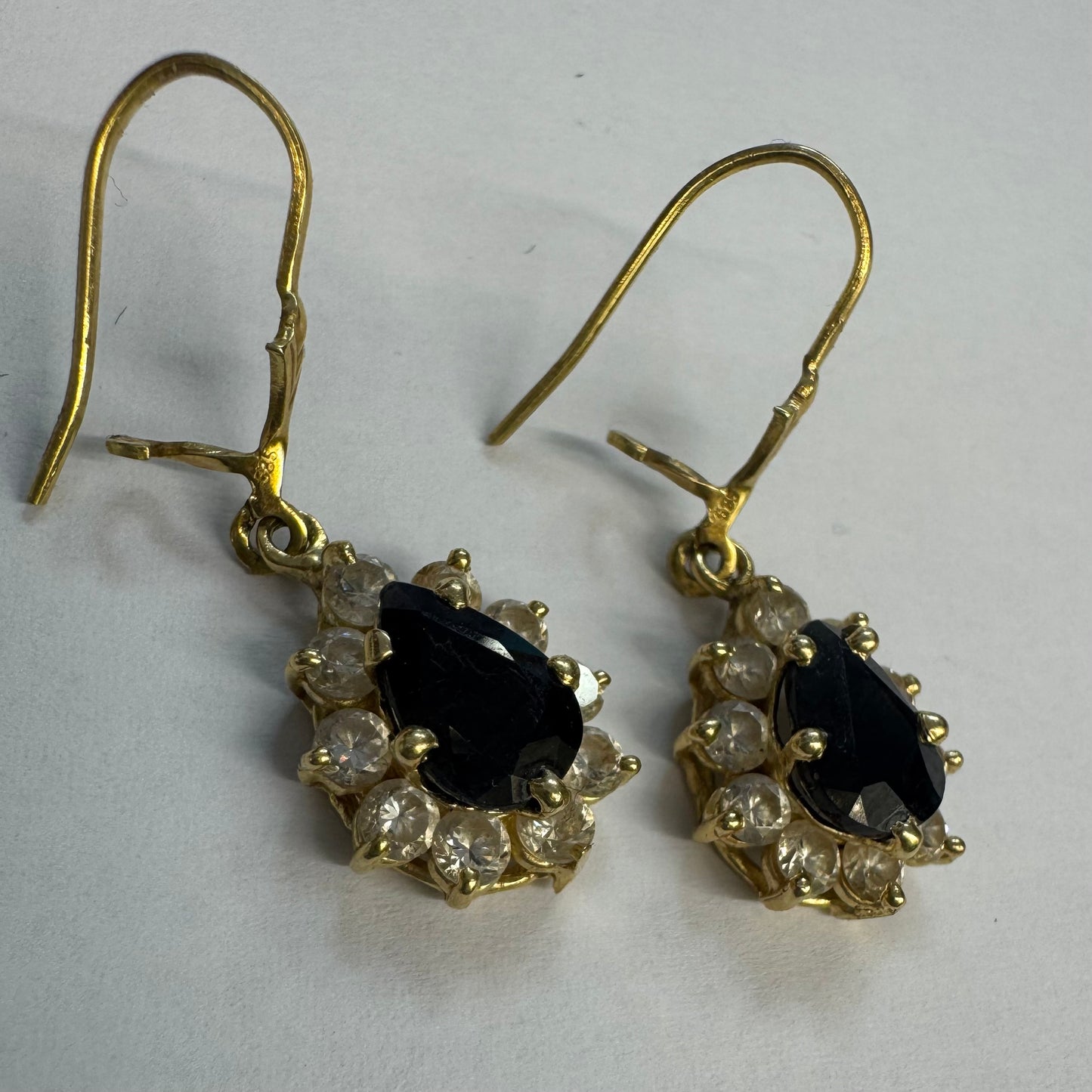 עגילי זהב צהוב 14 קראט בעיצוב קלאסי ומרשים, משובצים אבן ספיר בגוון כחול כהה בחיתוך טיפה ומסביבה אבנים שקופות עם הילה נוצצת שמוסיפה אור וזוהר לכל תנועה.