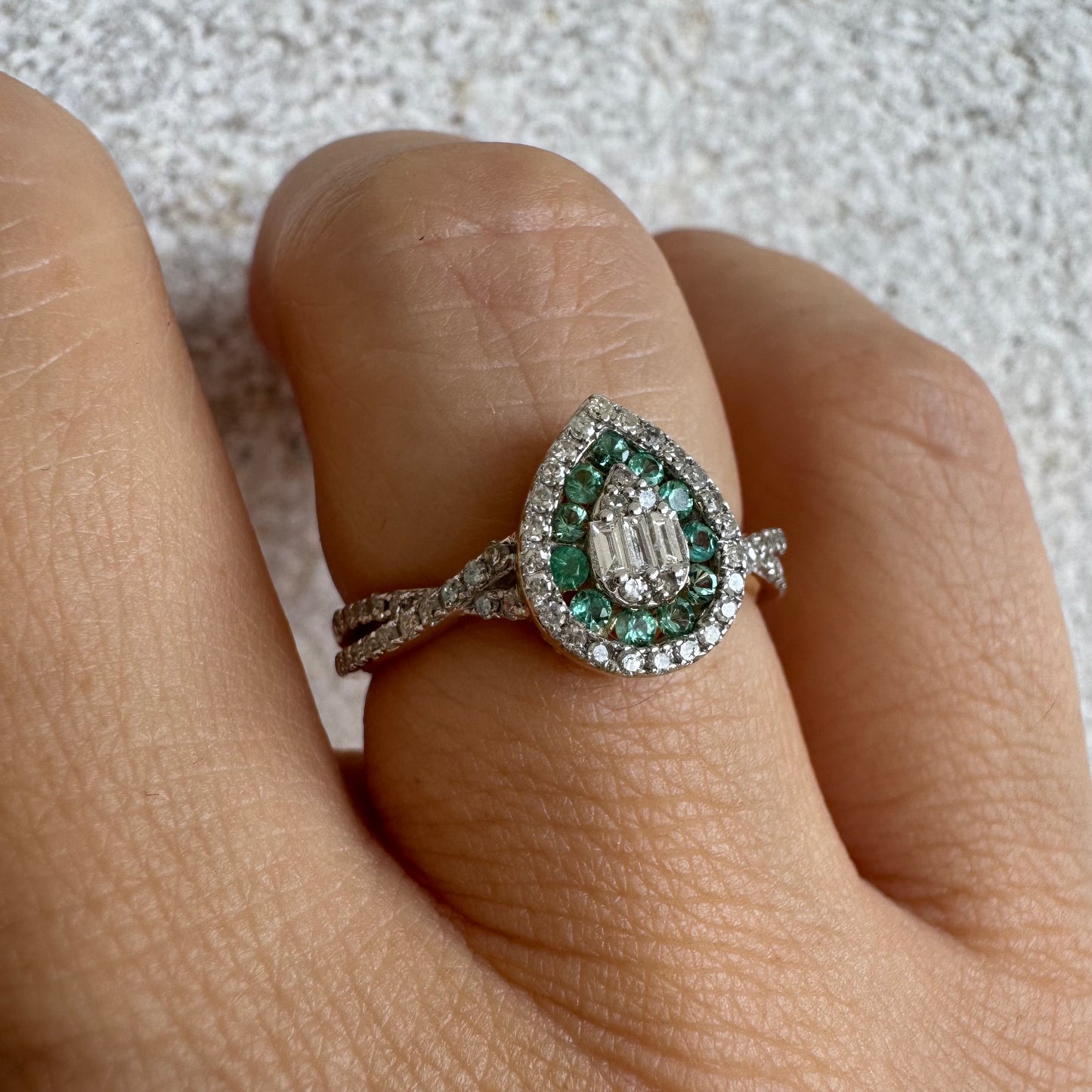 טבעת זהב צהוב 14 קראט בעיצוב טיפה (pear shape), משובצת יהלומים לבנים בשיבוץ הילה כפולה (double halo) ואבני אמרלד קטנות המקיפות מרכז ייחודי של יהלומים בחיתוך באגט