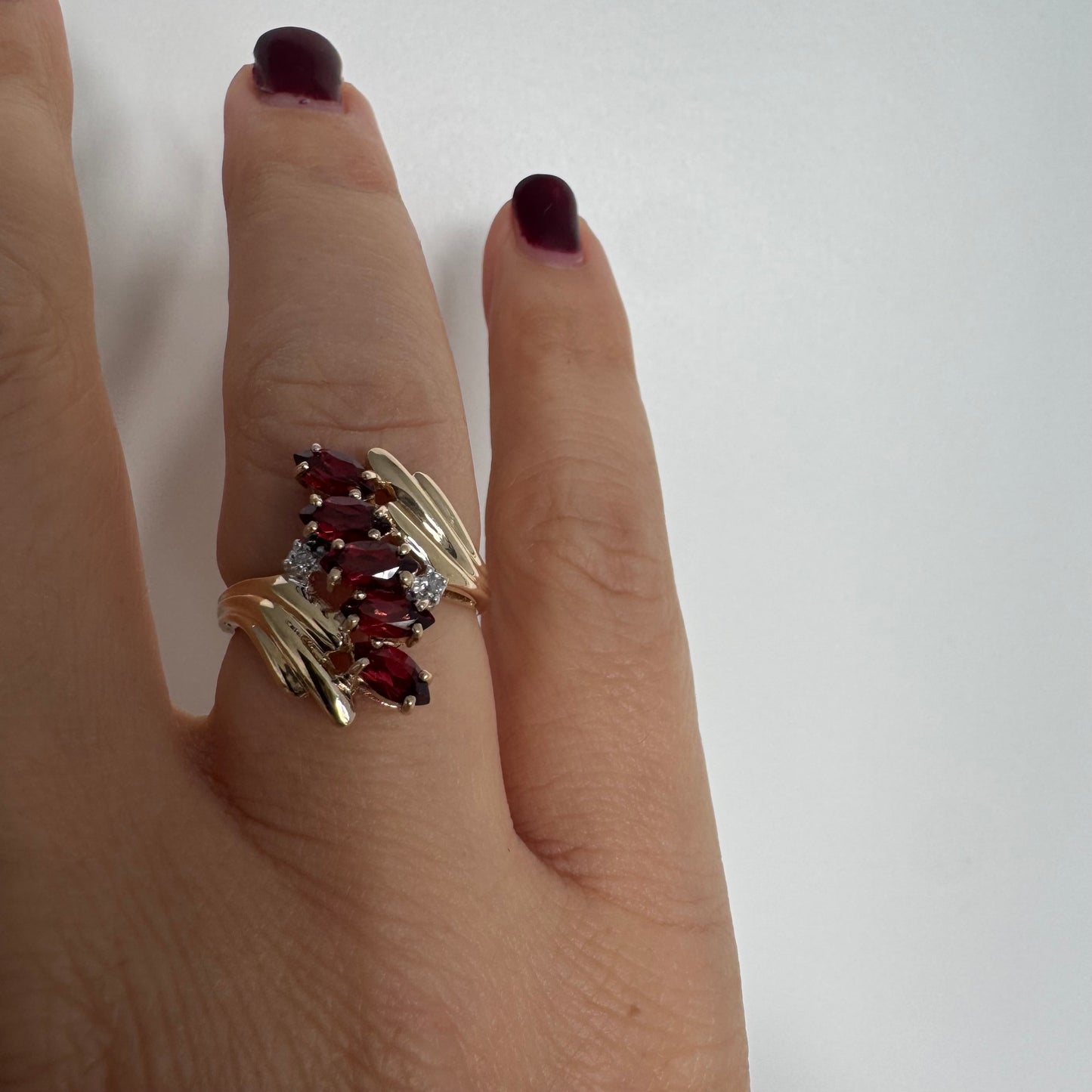 טבעת זהב צהוב 10 קראט בעיצוב דינמי ומלא תנועה, משובצת אבני גרנט בחיתוך מרקיזה בגוון אדום עמוק ובמרכזה נגיעות יהלומים לבנים.