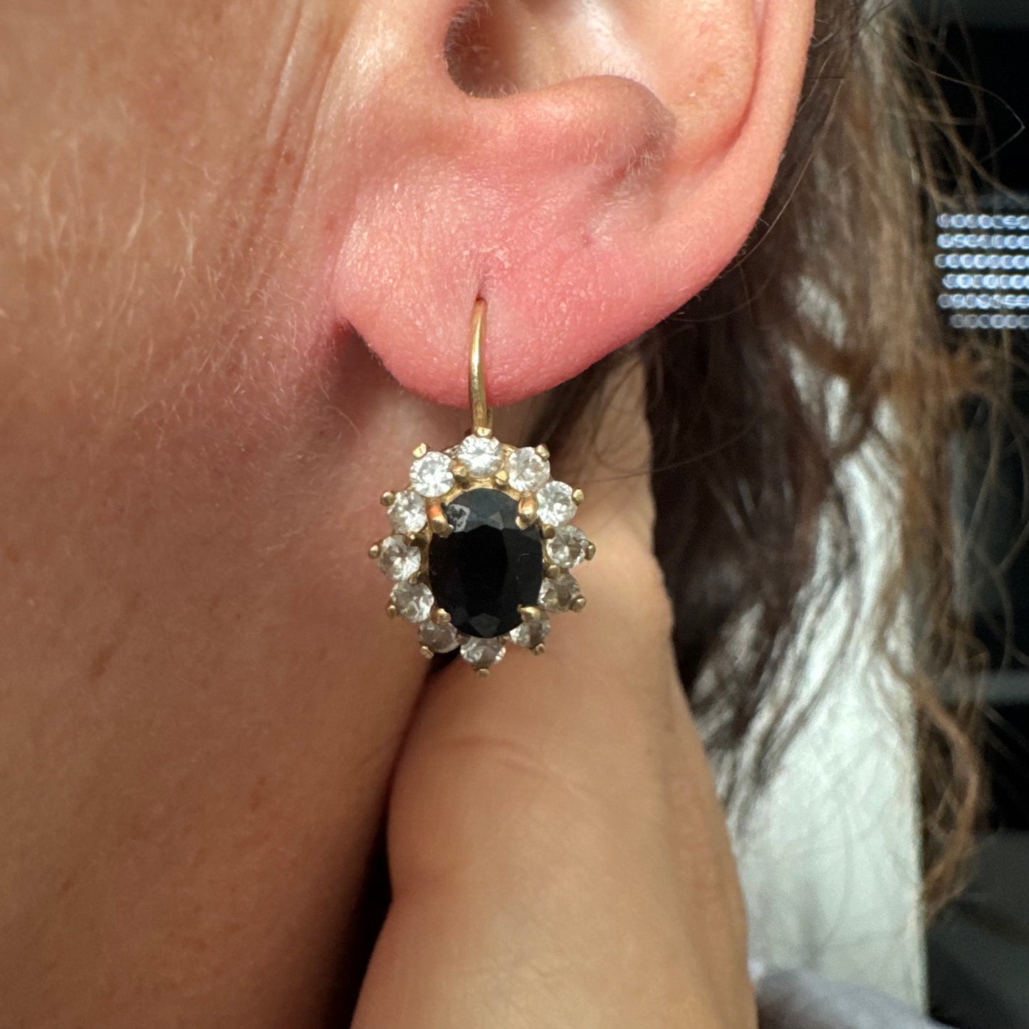 עגילי זהב צהוב 14 קראט בעיצוב קלאסי ומרשים, משובצים אבן ספיר בגוון כחול כהה בחיתוך אובלי, ומסביבה משובצים אבנים נוצצות שמוסיפות אור וזוהר לכל תנועה.