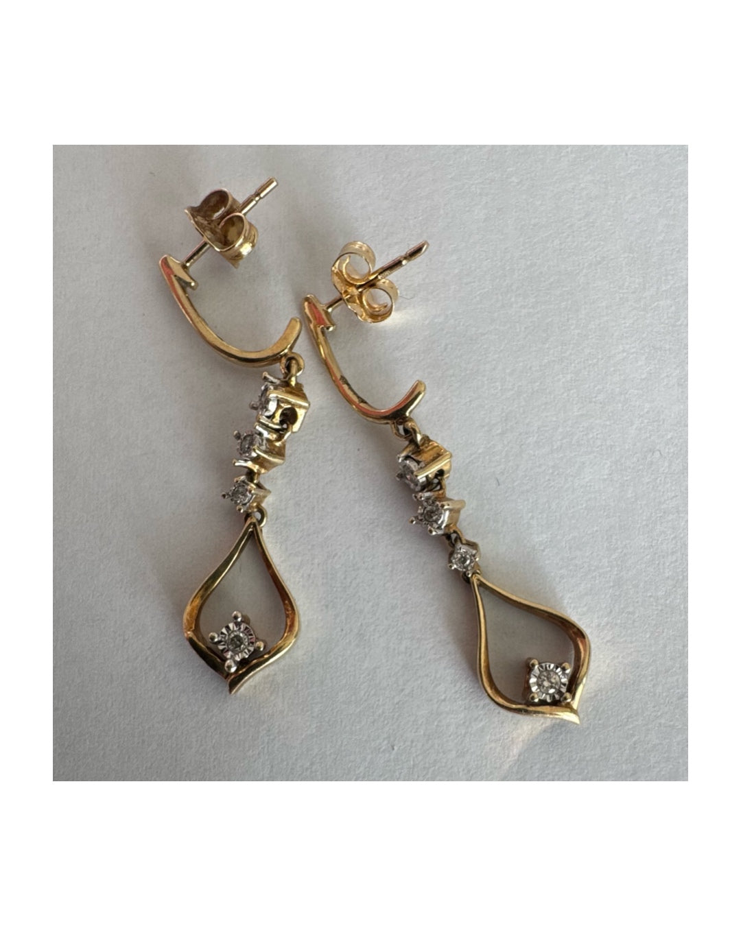 עגילי זהב 14 קראט תלויים בעיצוב עדין ומודרני, משובצים יהלומים לבנים לאורך החלק המרכזי ובטיפה פתוחה בתחתית משובץ יהלום נוסף.
