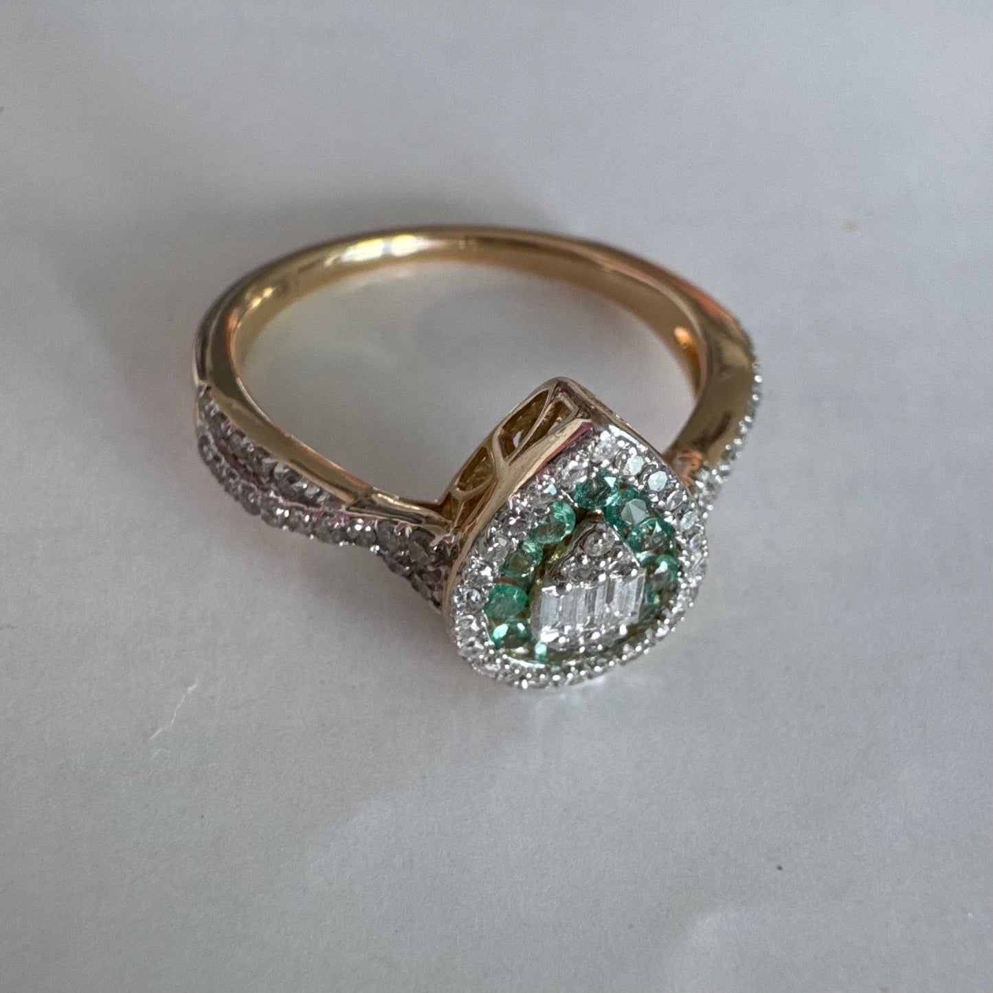 טבעת זהב צהוב 14 קראט בעיצוב טיפה (pear shape), משובצת יהלומים לבנים בשיבוץ הילה כפולה (double halo) ואבני אמרלד קטנות המקיפות מרכז ייחודי של יהלומים בחיתוך באגט